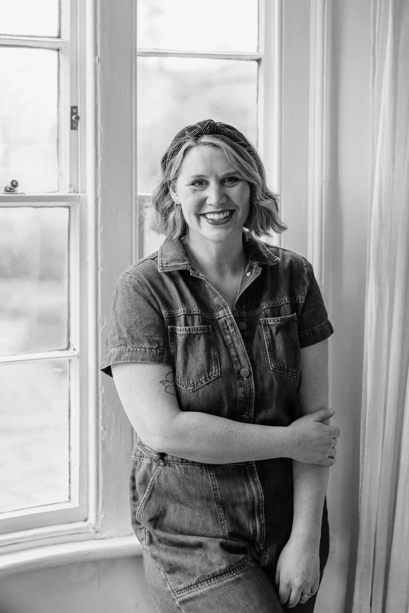 A smiling woman with shoulder-length wavy hair standing by a window in a casual denim outfit.