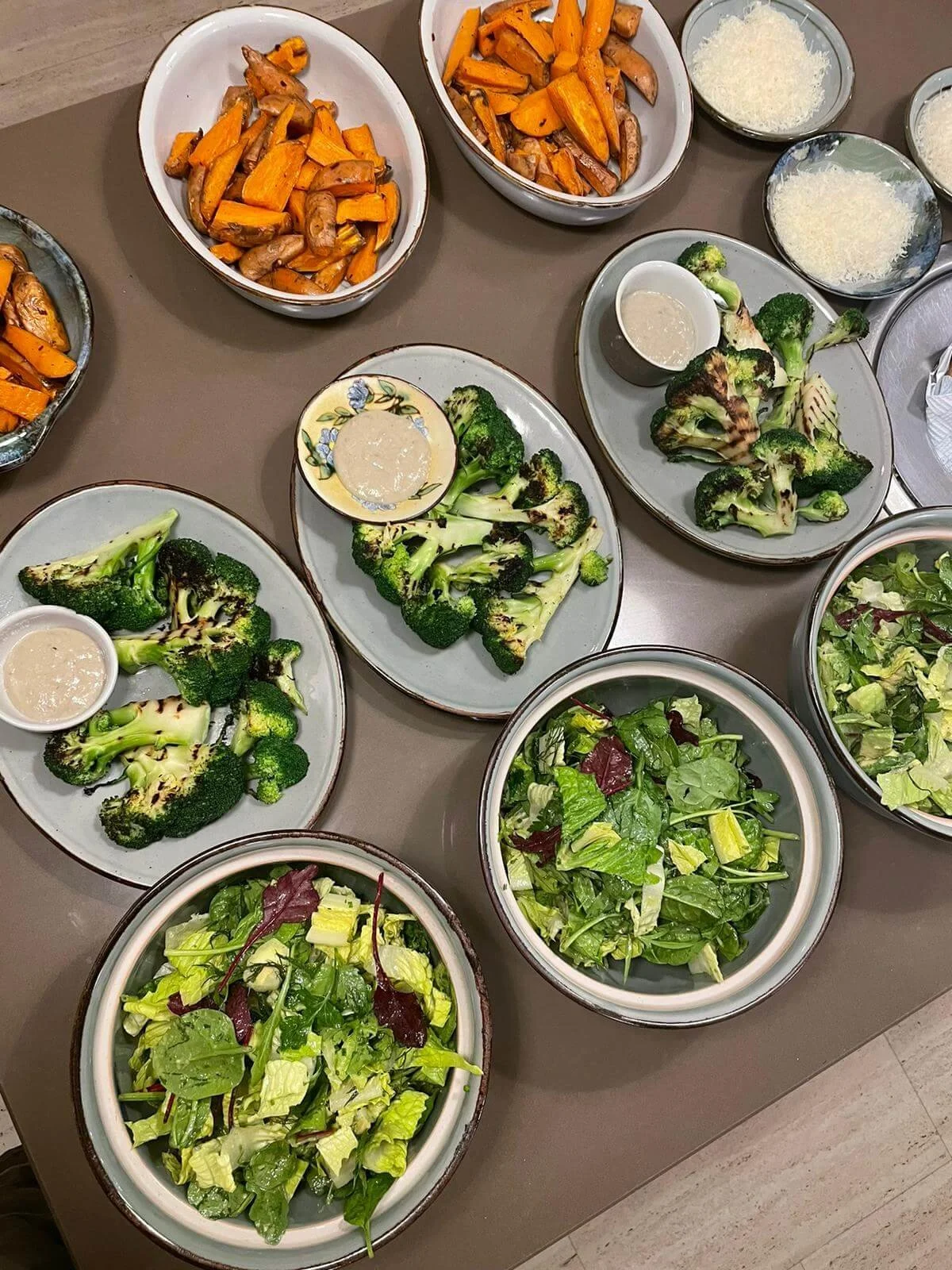 Assorted bowls of roasted sweet potatoes and mushrooms, bowls of shredded cheese, and plates of grilled broccoli and fresh mixed salads on a table.