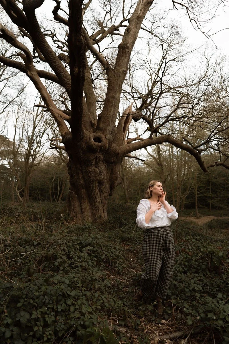A woman with shoulder-length hair, wearing a white blouse and checkered pants, stands in a forested area with bare trees and dense green foliage, touching her face thoughtfully.