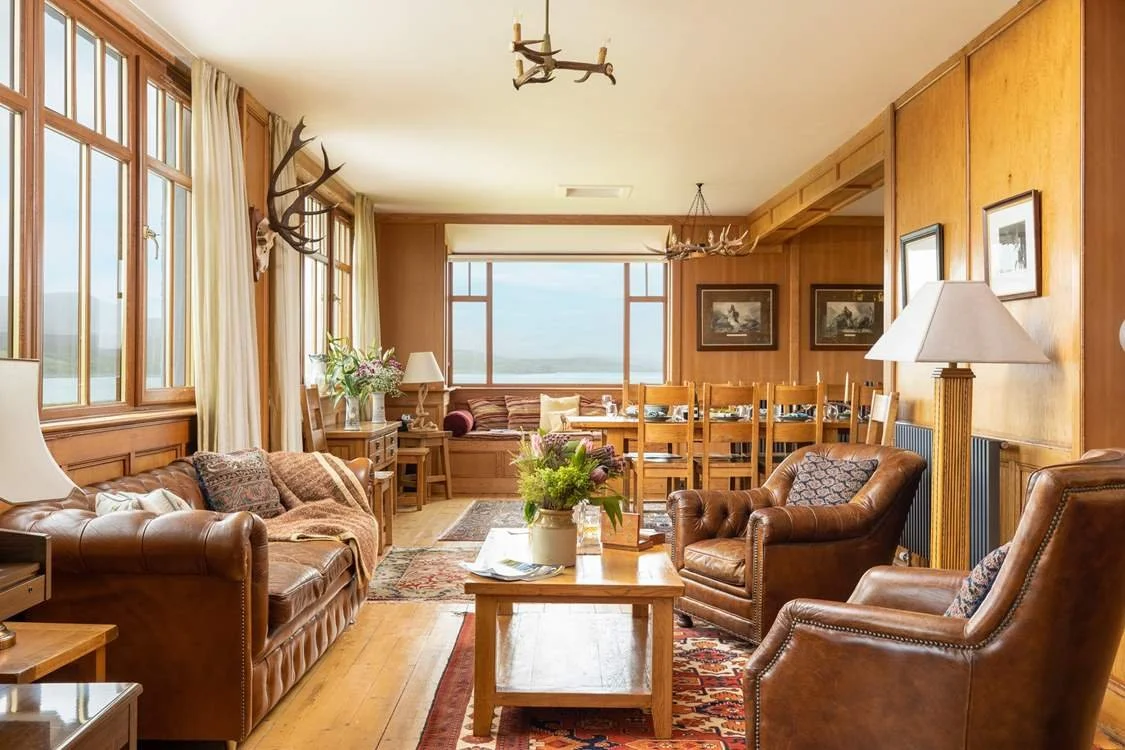 Living room with leather sofas, armchairs, wooden walls, large windows, and mountain views.