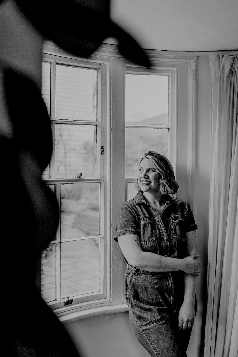 Black and white photo of a smiling woman with wavy hair, wearing a denim shirt and jeans, standing near a large window in a cozy room with curtains. Part of a person or object is visible in the foreground.