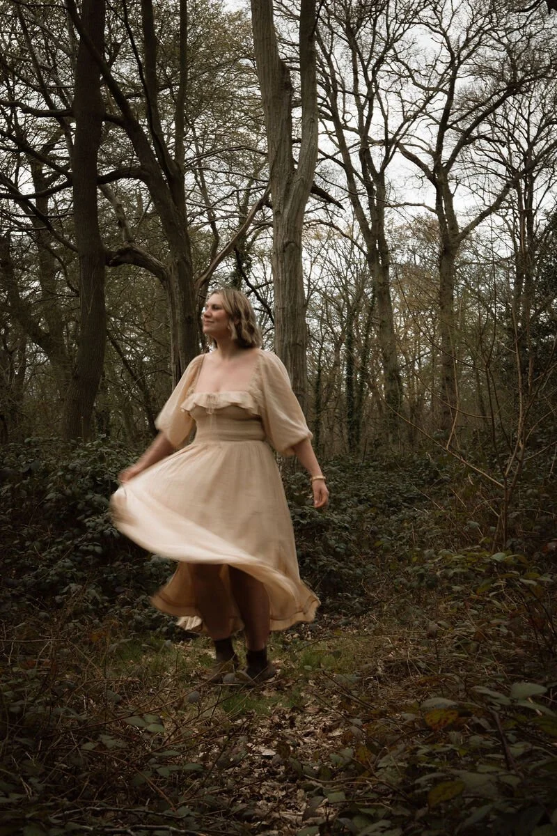 A woman in a flowing beige dress is spinning in a wooded forest with bare trees and dense foliage.