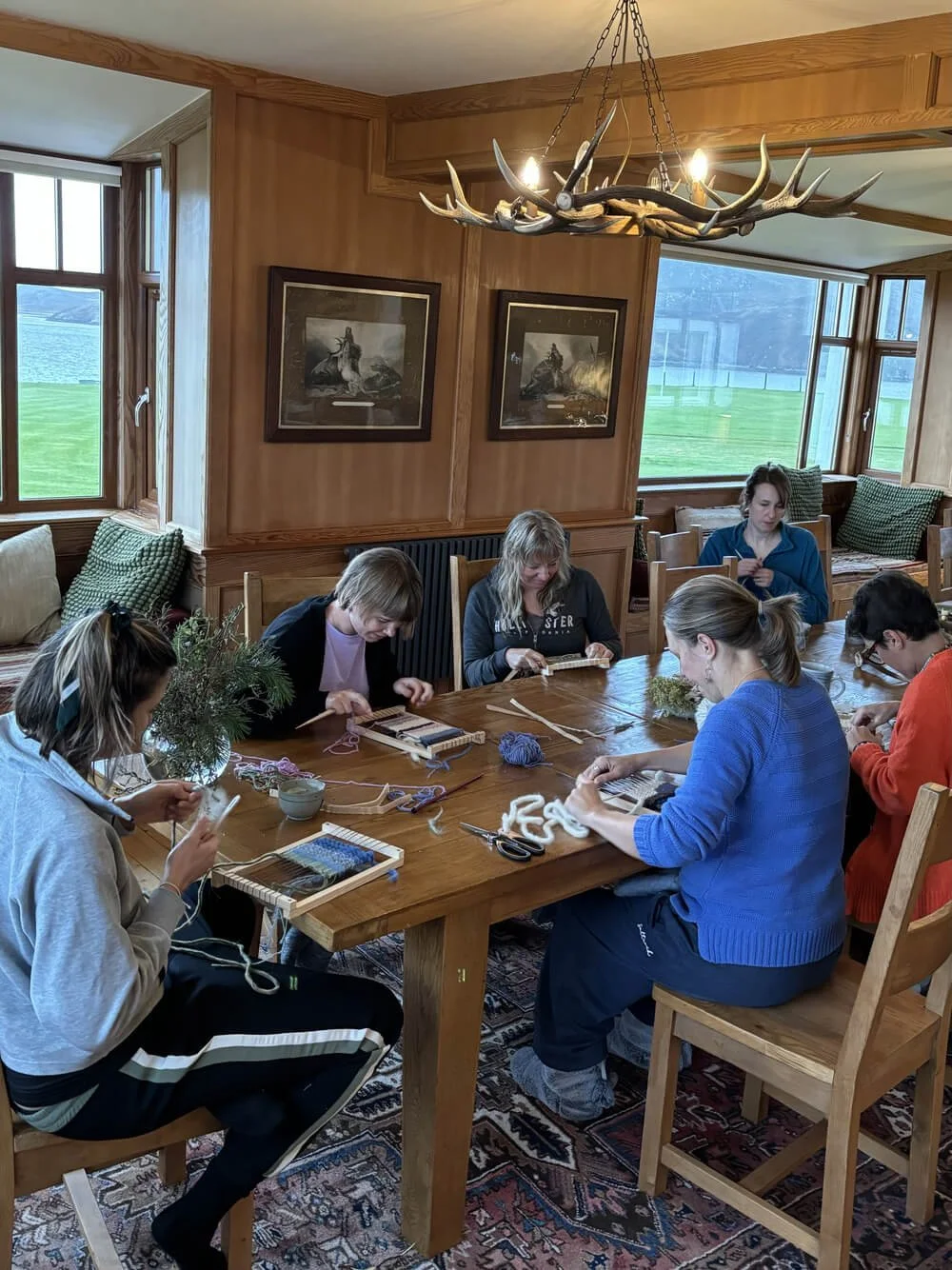Group of women sitting around a wooden table, knitting and working with yarn, in a cozy room with large windows and wooden walls.