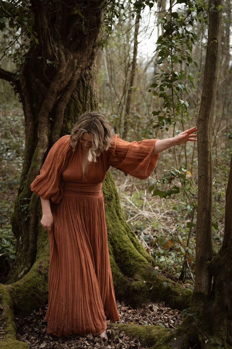 A woman in a long, pleated, rust-colored dress standing in a moss-covered forest. She is touching a tree with one hand and looking down, with her hair slightly tousled.