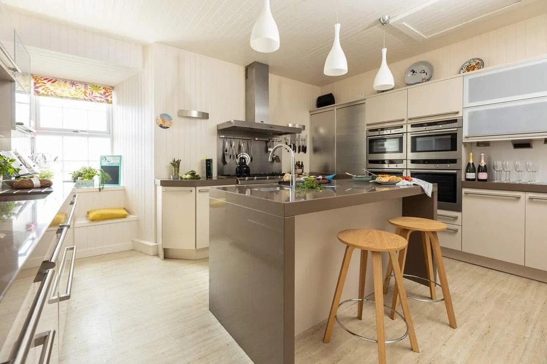 Modern kitchen with white cabinetry, stainless steel appliances, a kitchen island, two wooden bar stools, pendant lighting, and a window with a colorful valance.