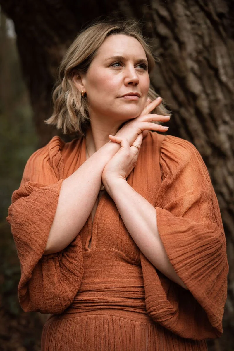 A woman with short blonde hair and fair skin is standing outdoors near a large tree, wearing an orange-brown dress with puffed sleeves. She is touching her neck and chin with her hands, looking thoughtfully into the distance.