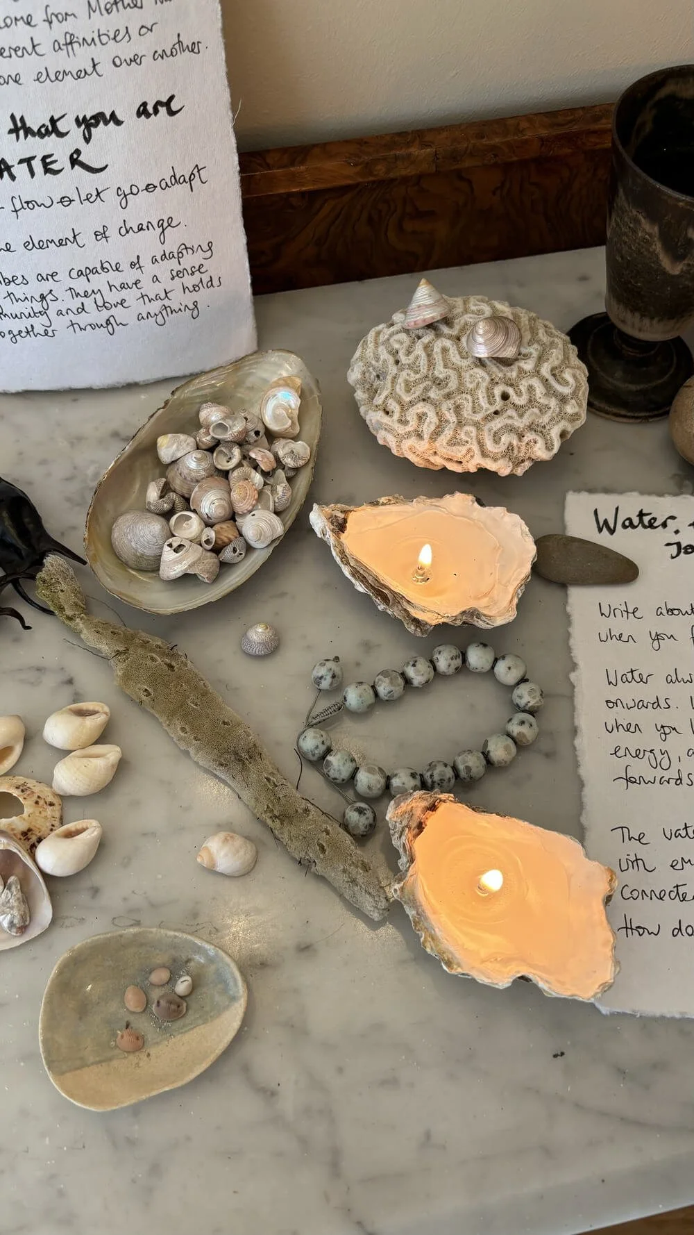 A collection of seashells, coral, candles, stones, and handwritten notes on a marble surface, with a wooden trim in the background.