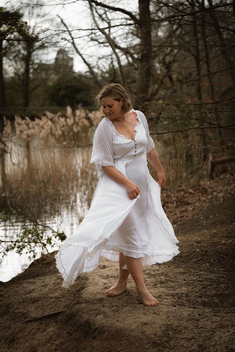 A woman in a white dress standing on a dirt path next to a body of water with trees in the background.