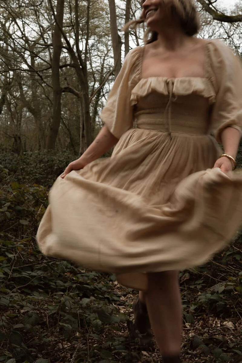 A smiling woman in a beige dress walking through a wooded area, holding the hem of her dress while moving.