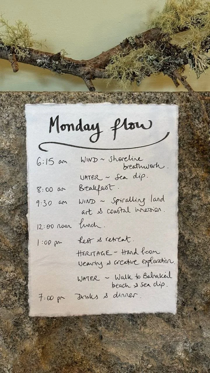 A handwritten daily schedule on a piece of paper, listing activities from 6:15 am to 7:00 pm, including wind, water, breakfast, art, lunch, rest, exploration, walk to beach, and dinner, with a decorative branch above and a granite countertop below.