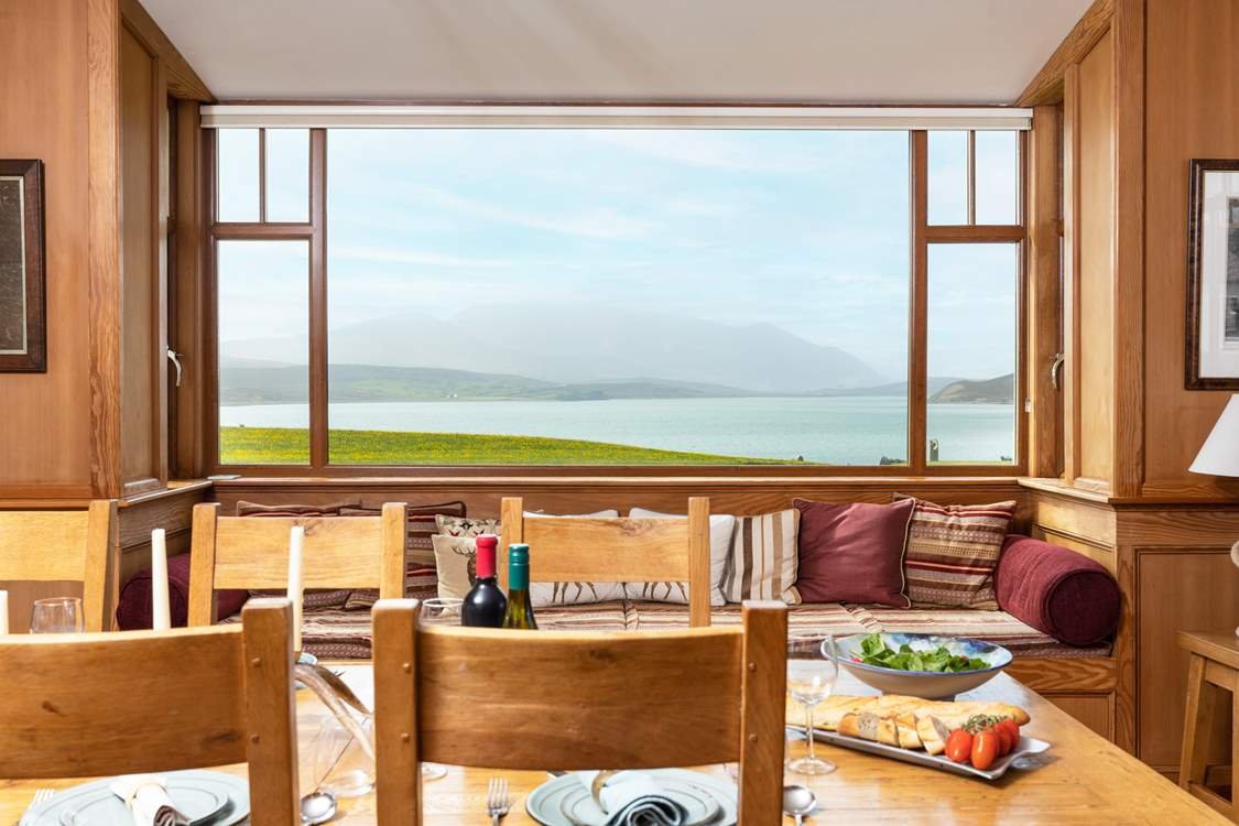 Dining room with a large window view of a lake and mountains, wooden furniture, and a table set with food and drinks.