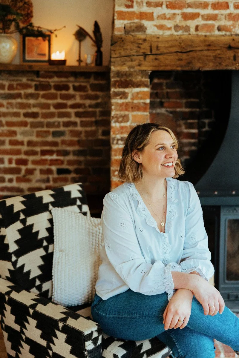 A woman with shoulder-length wavy hair sitting on a black and white patterned couch, smiling and looking to the right. She is wearing a white embroidered blouse and blue jeans. The background features a brick fireplace with a wooden mantel decorated with a candle, photo frame, and small decorative items, creating a cozy home interior.