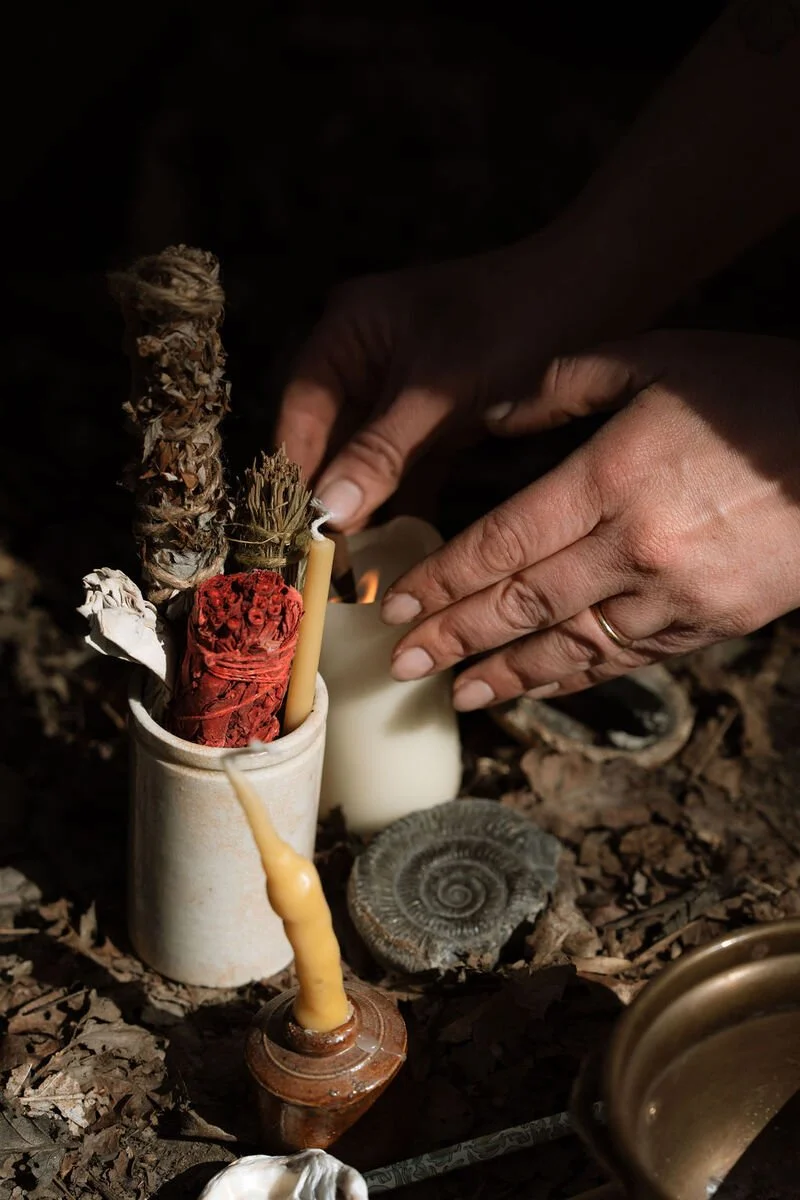Person lighting a candle on a small altar with dried herbs, seashells, and other objects.