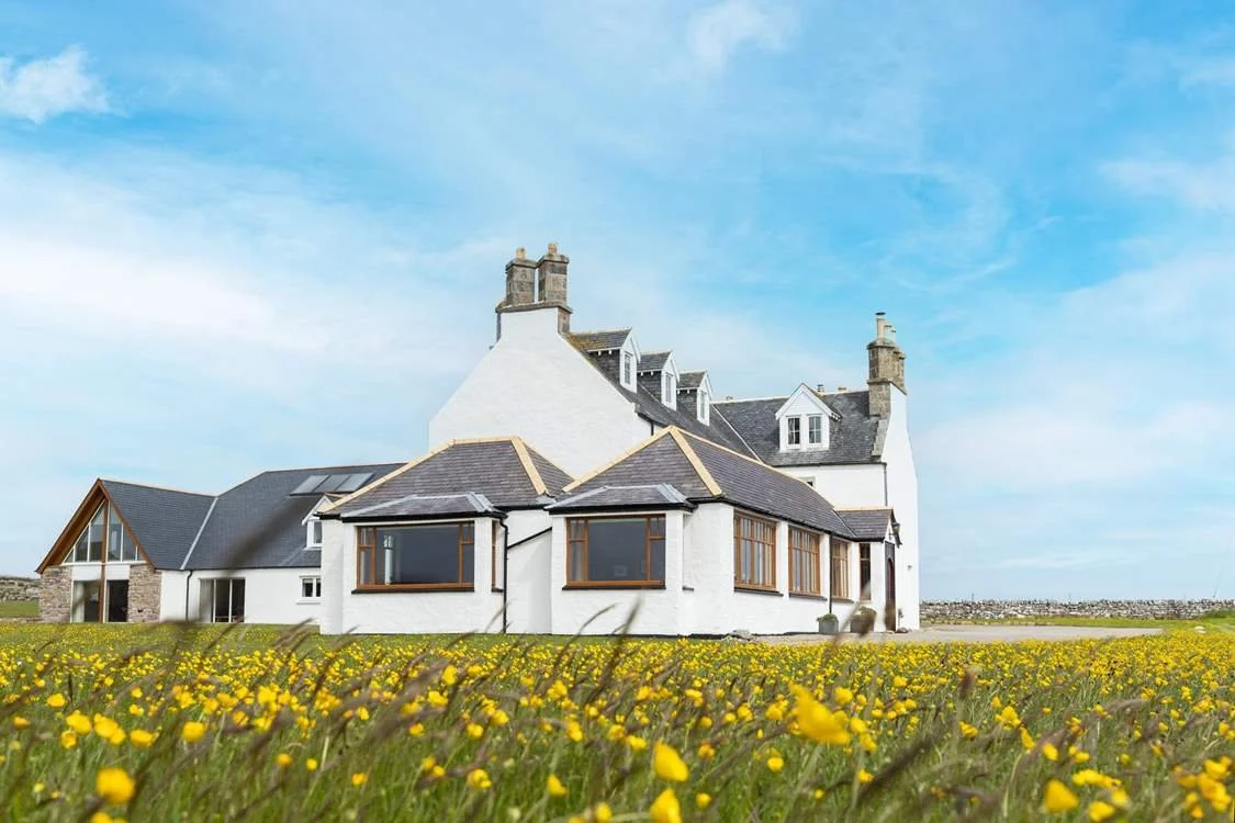 A large white house with a mix of flat and gabled roofs, surrounded by a field of yellow flowers under a blue sky with light clouds.