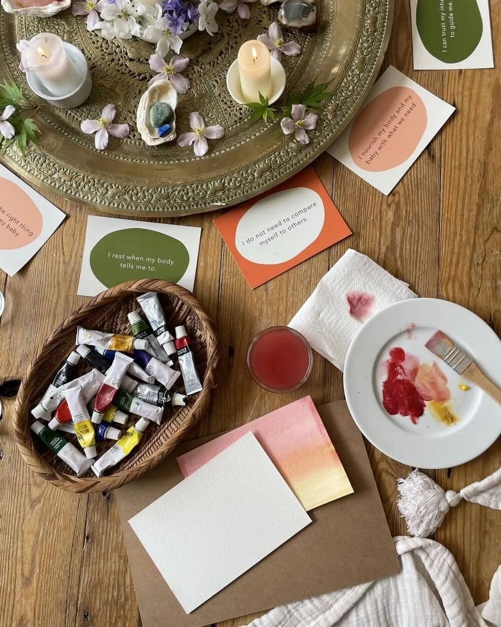 Table with candles, flowers, seashells, cards with affirmations, a basket of paint tubes, a plate with paint, a glass of red liquid, and notecards.