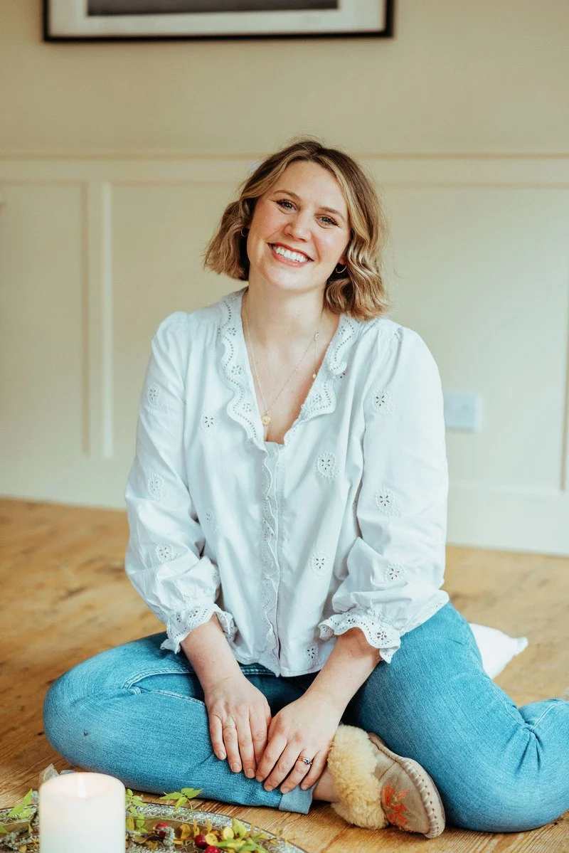 A woman sitting cross-legged on the floor, smiling, wearing a white blouse and blue jeans, with a candle and decorative items in front of her.