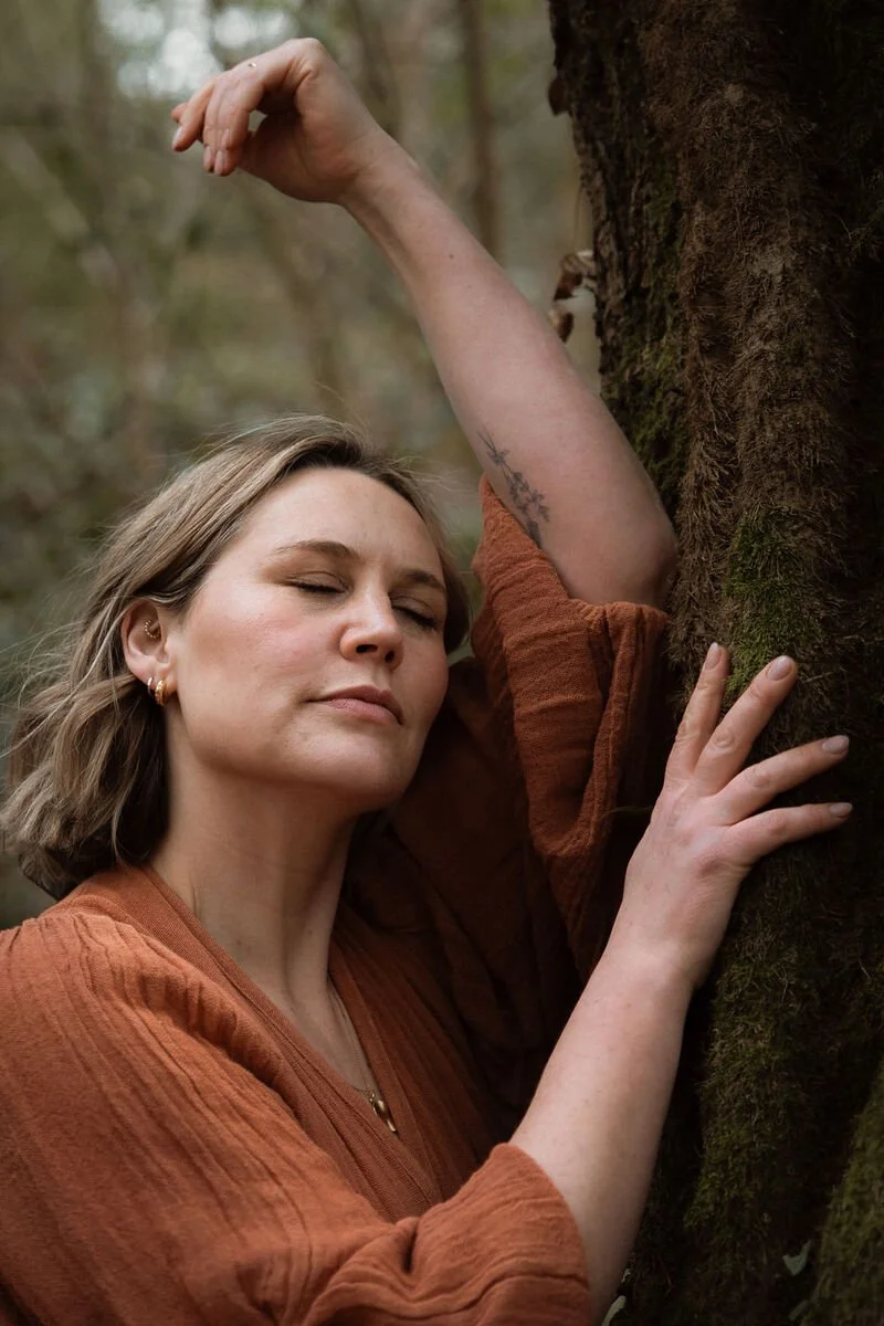 A woman with closed eyes gently hugging a tree in a forest during daytime, appearing peaceful and content.
