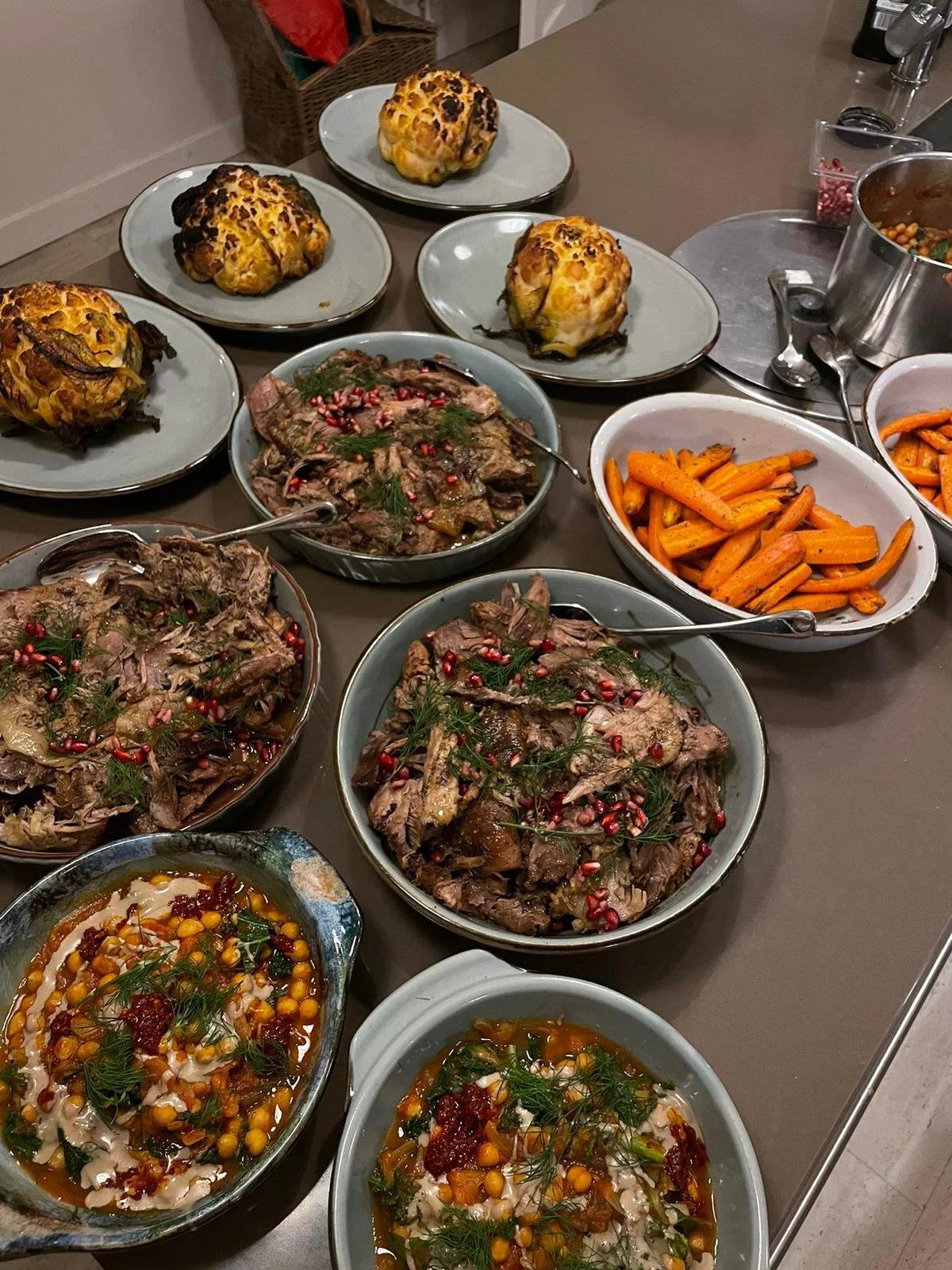 A table filled with various dishes including roasted vegetables, pulled meat with garnishes, and baked bread rolls, set in serving bowls and plates.