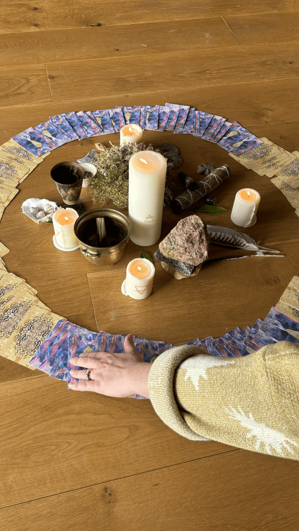 A circular arrangement of candles, crystals, feathers, and tarot cards on a wooden table, with a person's hand resting on the edge of the table.