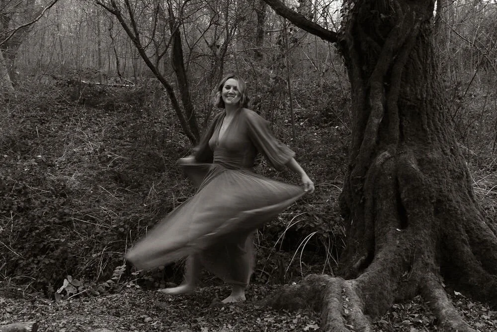 A woman in a long dress spinning and smiling outdoors near a large tree with bare branches.