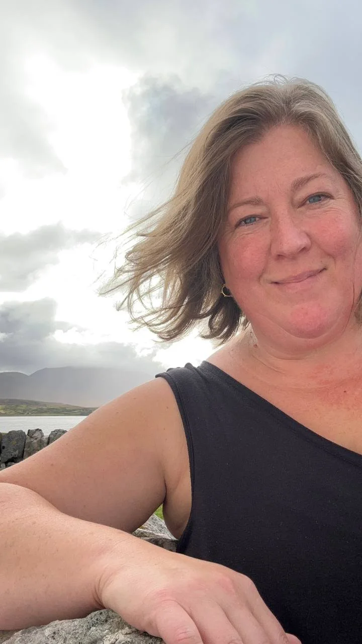 A woman with shoulder-length blonde hair and blue eyes, wearing a black sleeveless top, smiling slightly at the camera with a cloudy sky and distant mountains in the background.