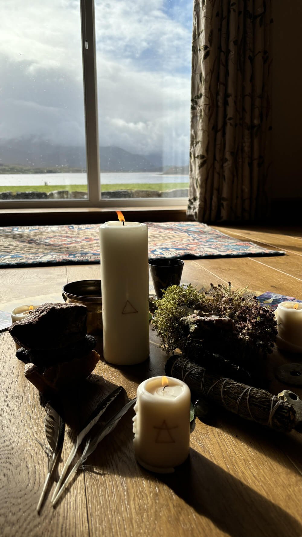 Decorative table setup with candles, stones, feathers, and herbs near a window with a lakeside view and mountains