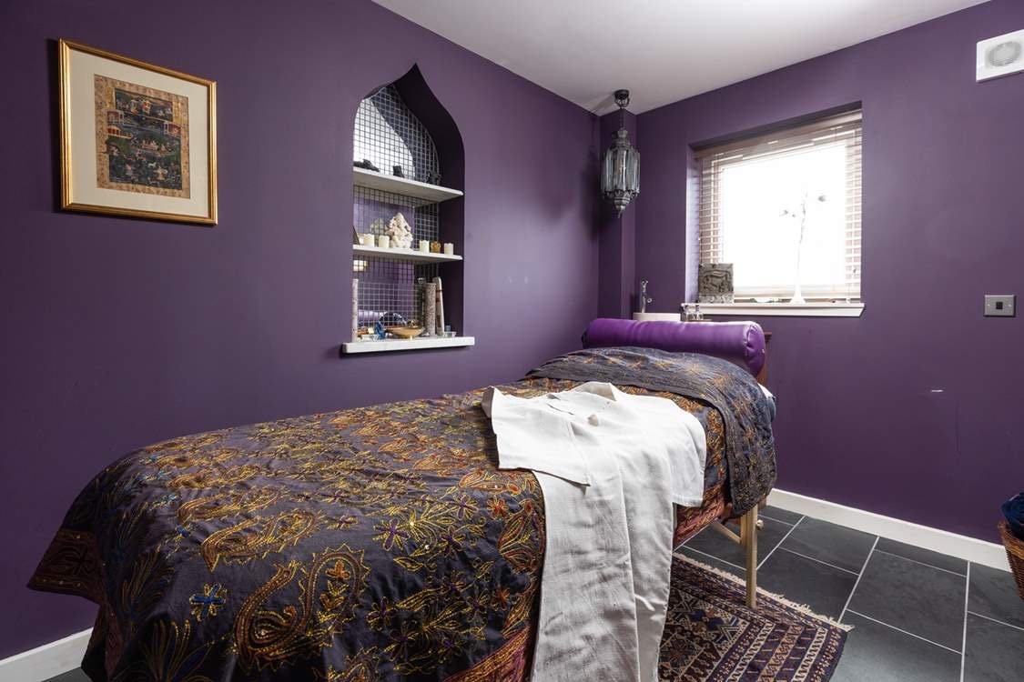 A purple-walled room with a massage table covered in an ornate, dark-colored blanket, and a white cloth draped over one side. A small window with blinds is on the right, and a niche with shelves containing various decorative items is in the background. A framed picture hangs on the left wall, and a hanging lantern-style light fixture is near the corner.