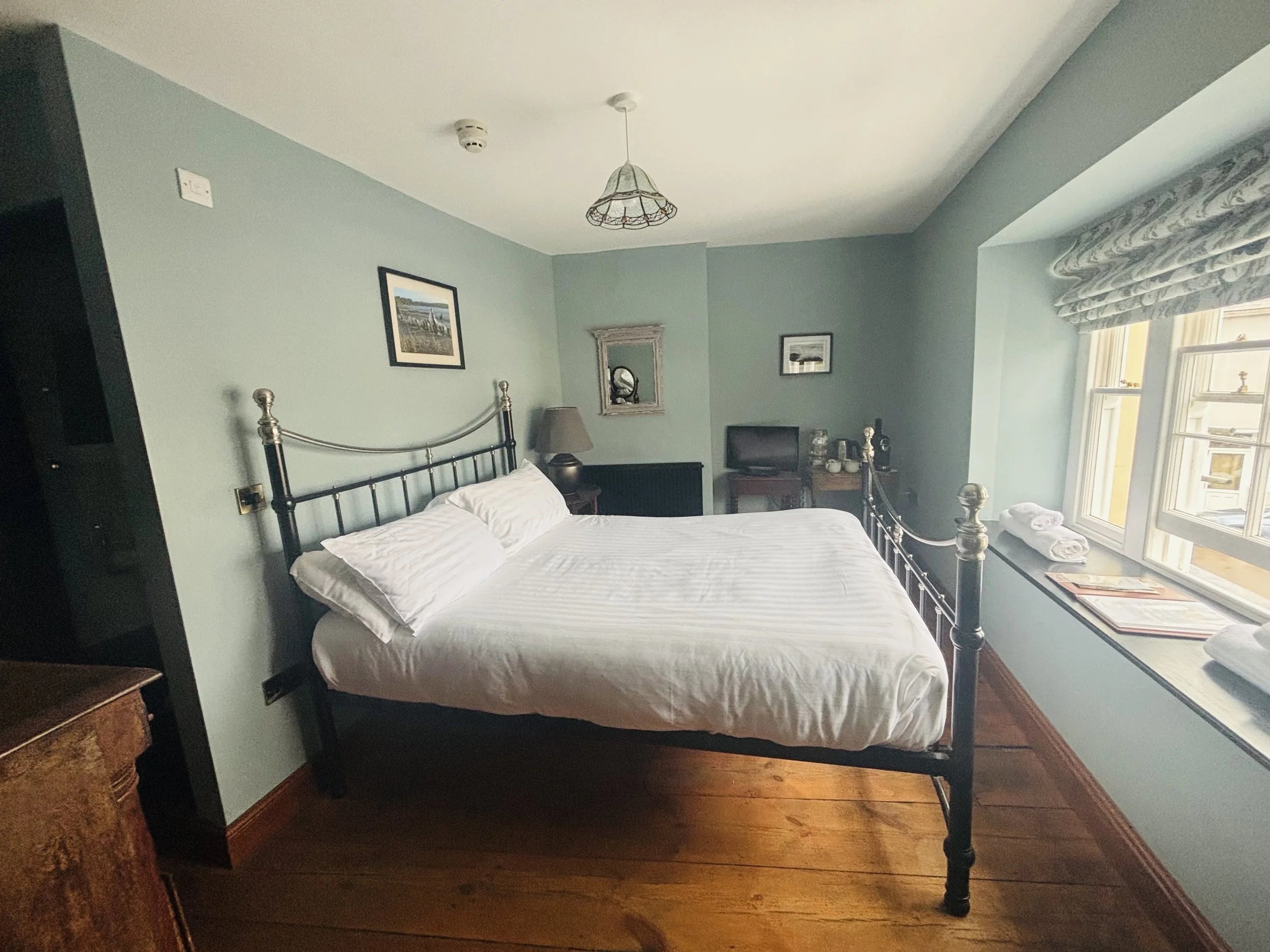 A cozy hotel room featuring a large bed with white linens and pillows, a small bedside table with a lamp, a window with patterned curtains, a wooden ledge with towels, and a small TV on a stand in the corner.