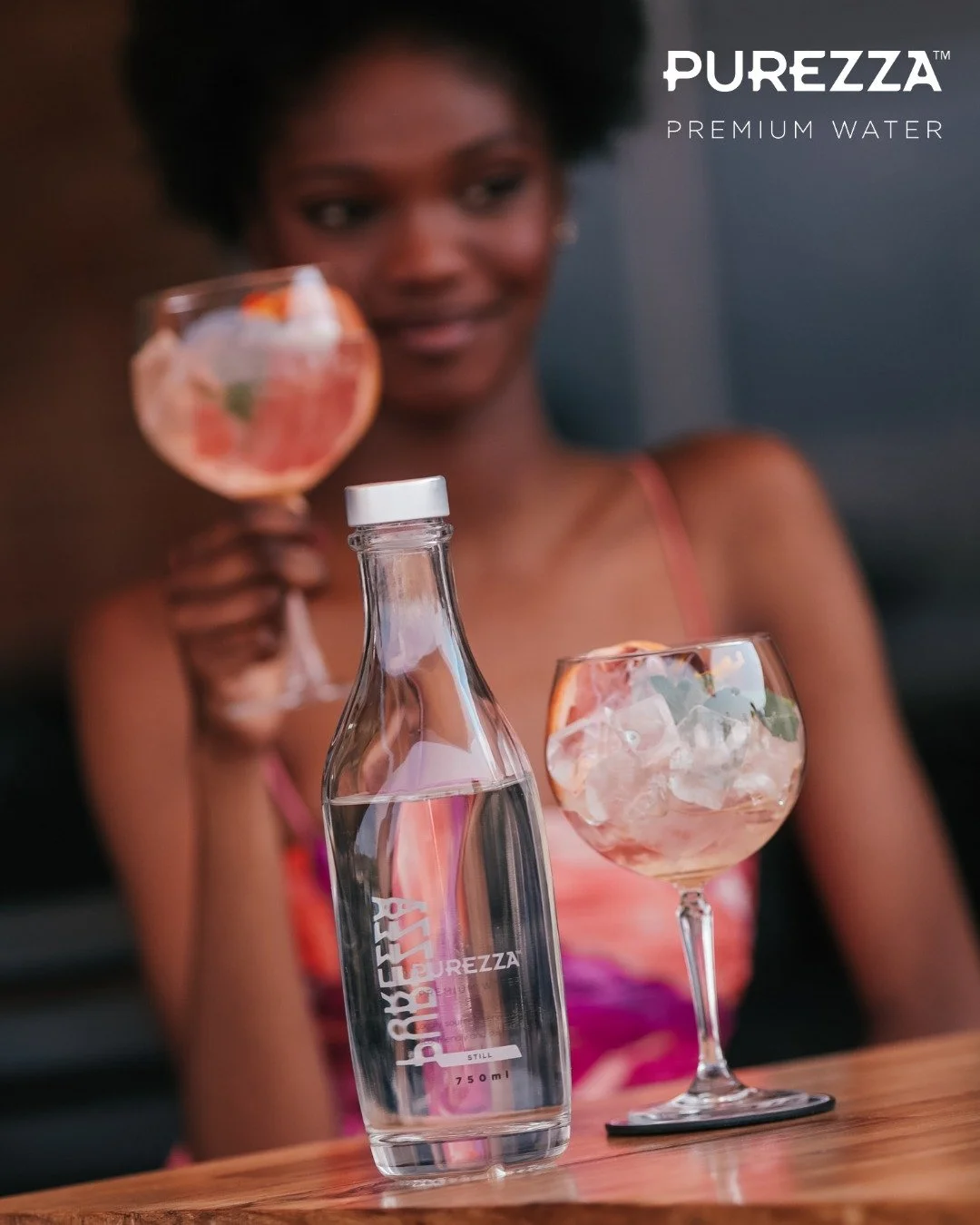 A woman holding a cocktail glass with ice, fruit, and mint, with a bottle of PUREZZA water and another cocktail glass on a wooden table.