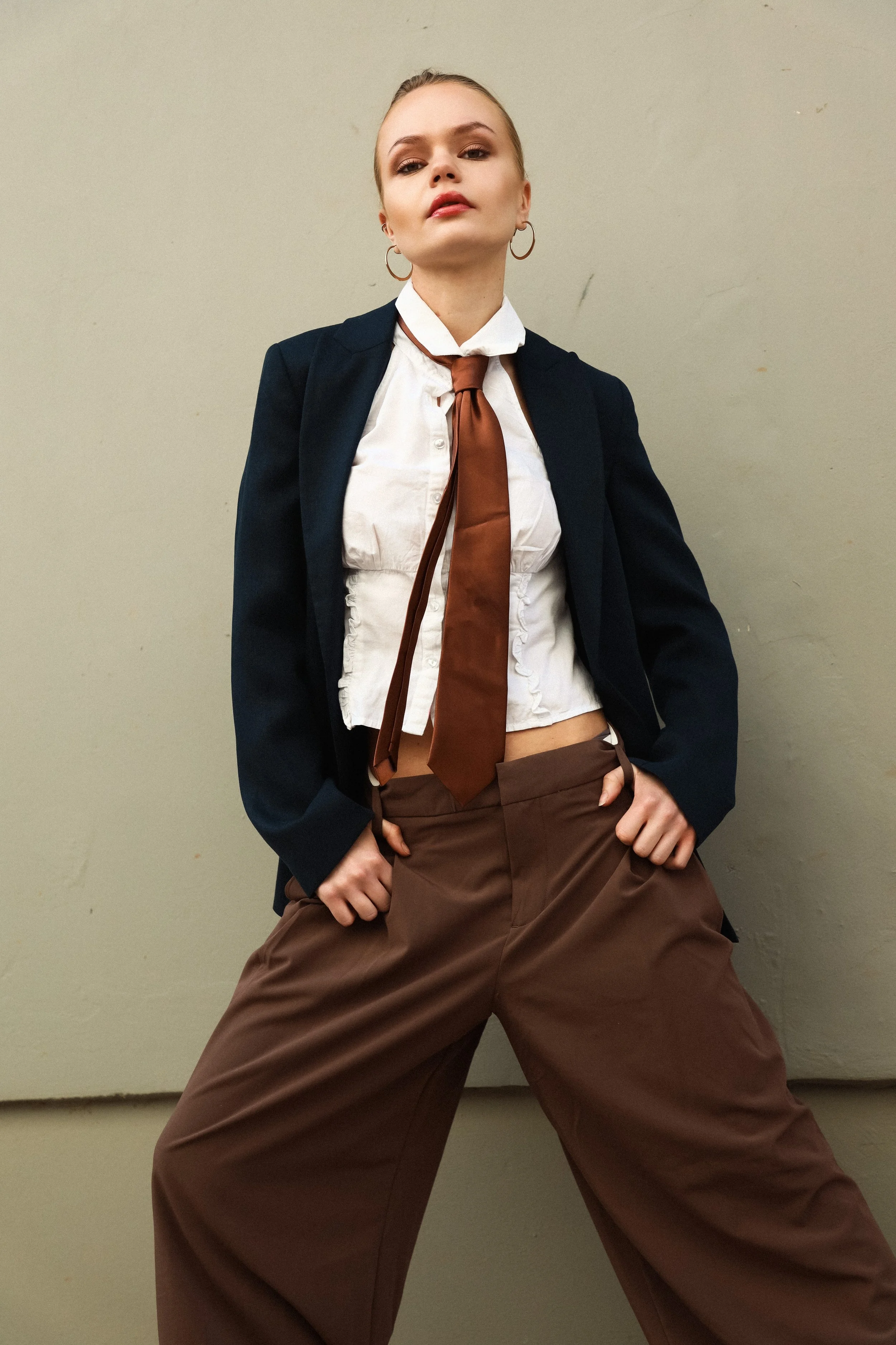 A woman wearing a black blazer, white shirt, brown tie, and brown pants, standing against a plain beige wall with her hands on her hips in a confident pose.