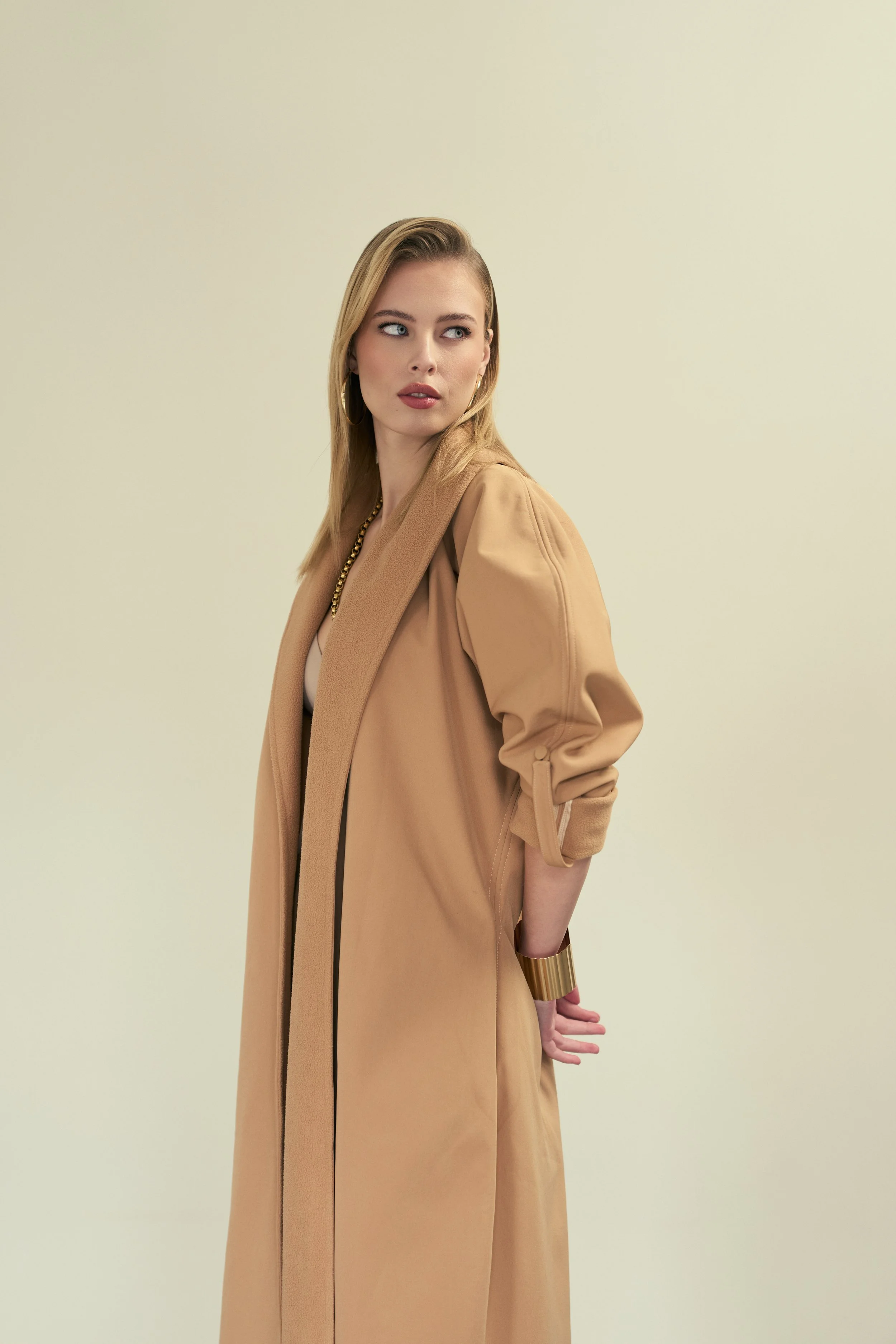 GAIA Fernandez from AURA Models wearing a tan trench coat, gold hoop earrings, and a gold cuff bracelet, standing against a plain light-colored background, looking to her right with a thoughtful expression.