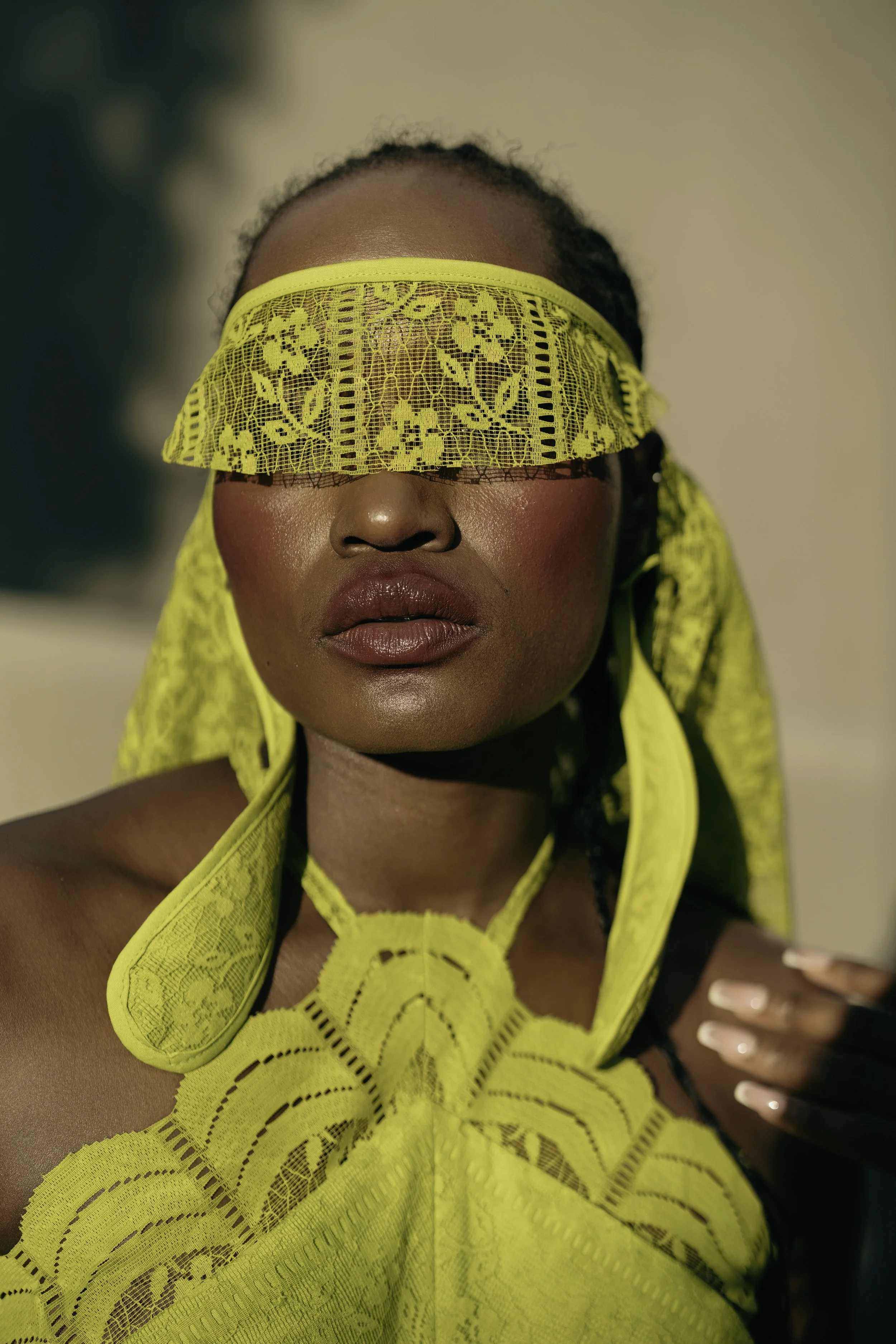 SHALOM Musvosvi from AURA Models with dark skin and full lips, wearing a yellow lace and mesh outfit that covers her eyes with a band of the same material, posing indoors against a light background.