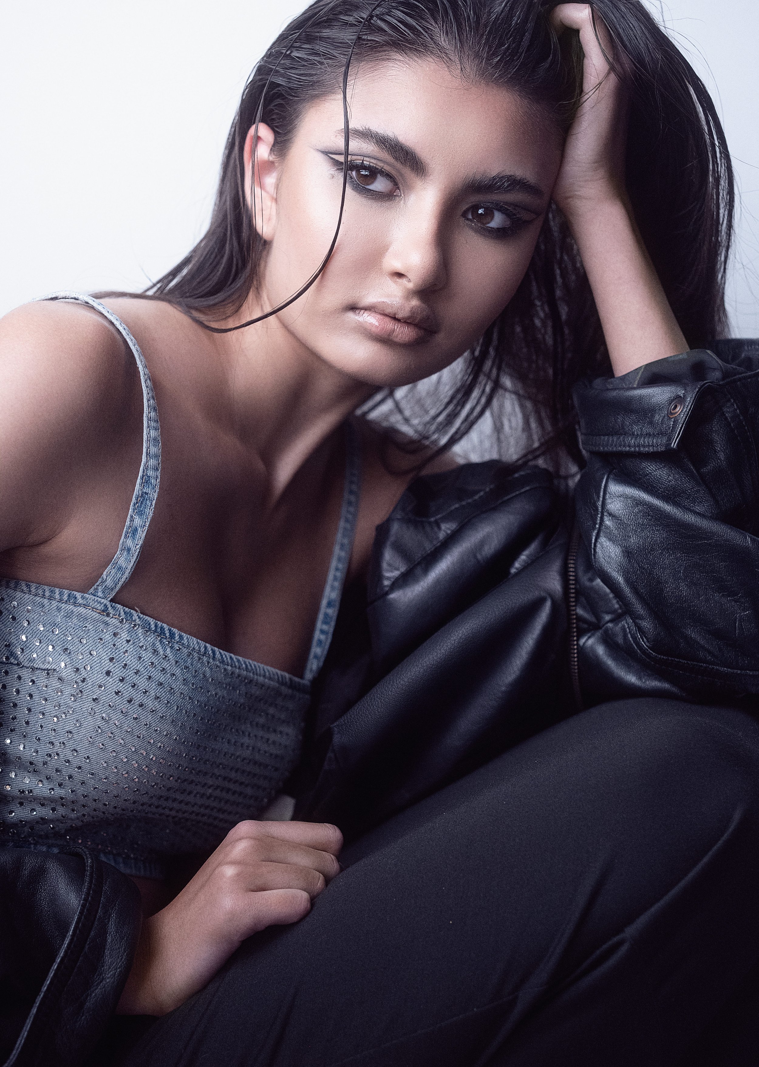 KEIRA Louw from AURA Models with long dark hair, wearing a gray, sparkly top and black leather jacket, looking to the side with her hand resting on her head, sitting in a relaxed pose against a plain background.