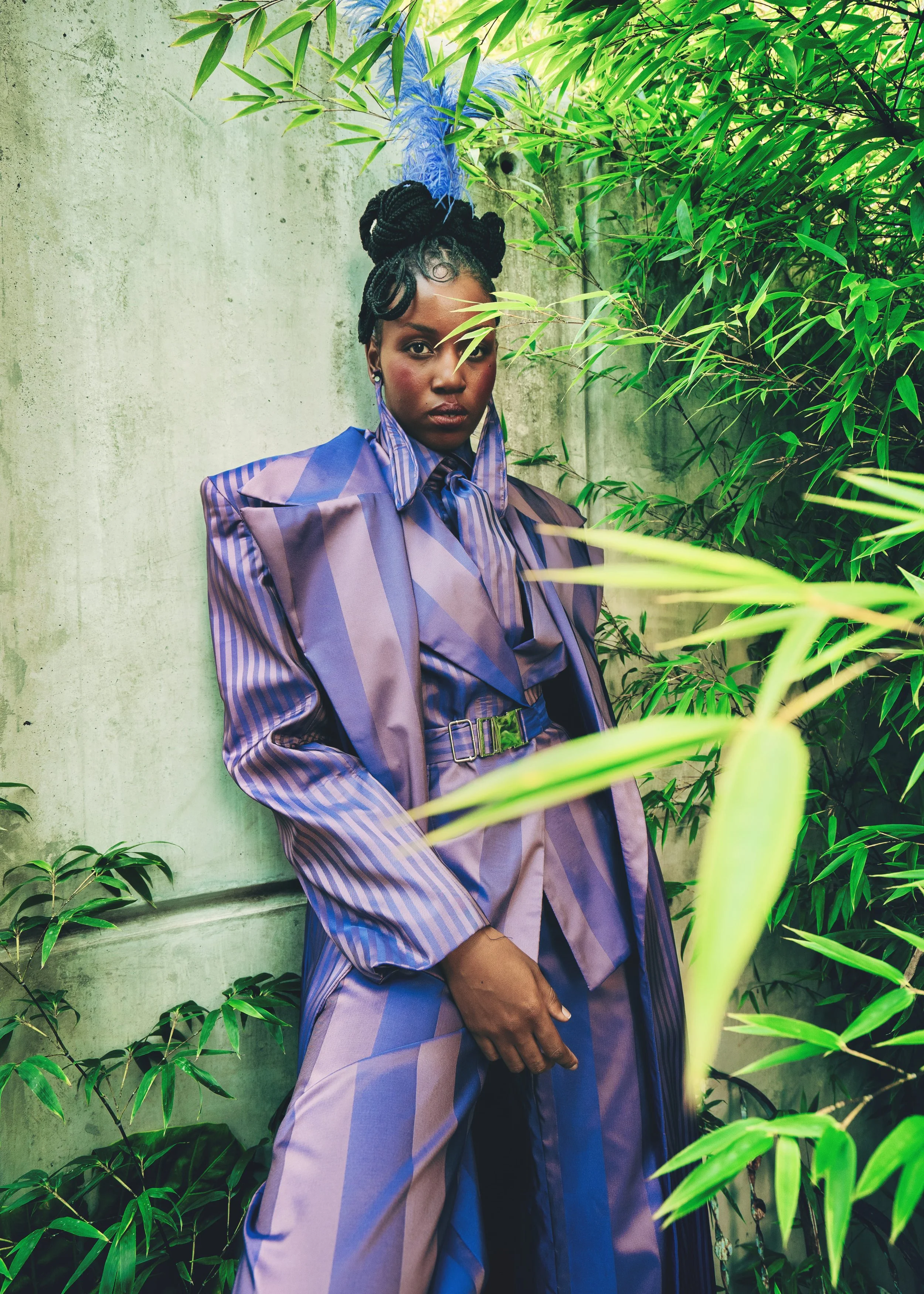 RIVO Baloyi from AURA Models with braided hair, dark skin, wearing a purple and blue striped outfit, standing against a concrete wall surrounded by green plants and foliage.