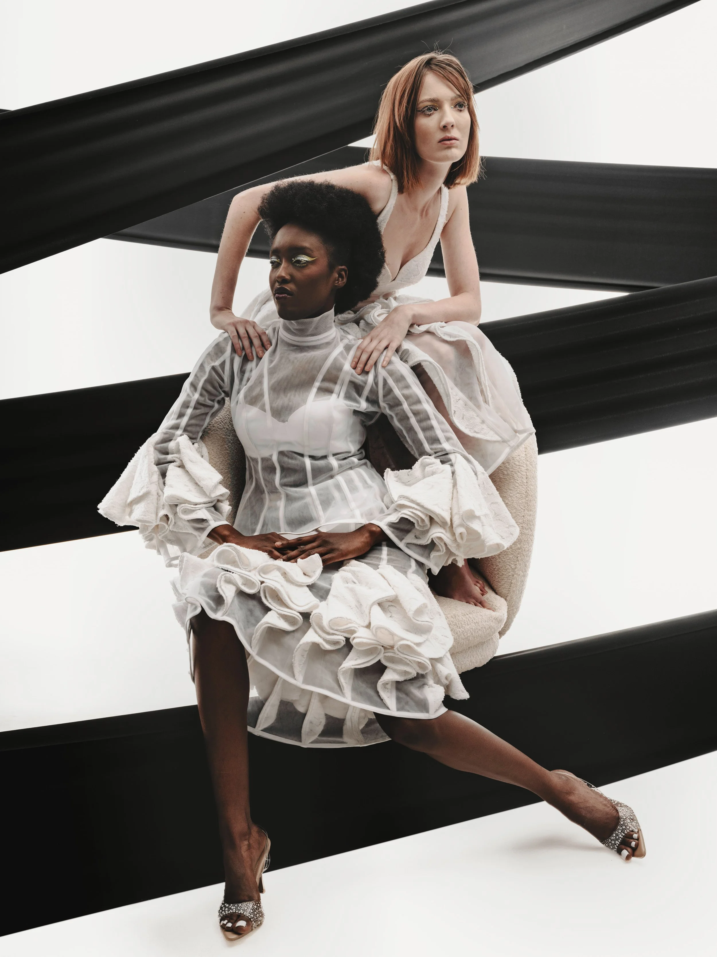 CAYLEIGH Bancroft and KIM Chakanyuka from AURA Models with contrasting hair styles and outfits seated on a beige chair against a white background with black angled beams. 