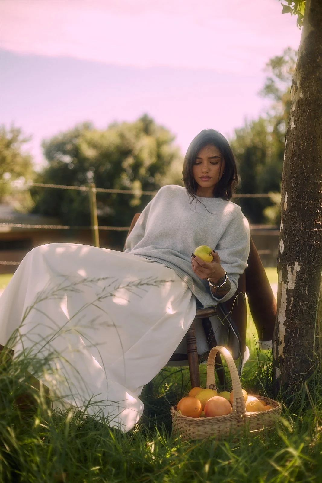 EMILY Francis from AURA Models sitting outdoors under a tree, holding an apple, with a basket of apples and pears on the grass beside her.