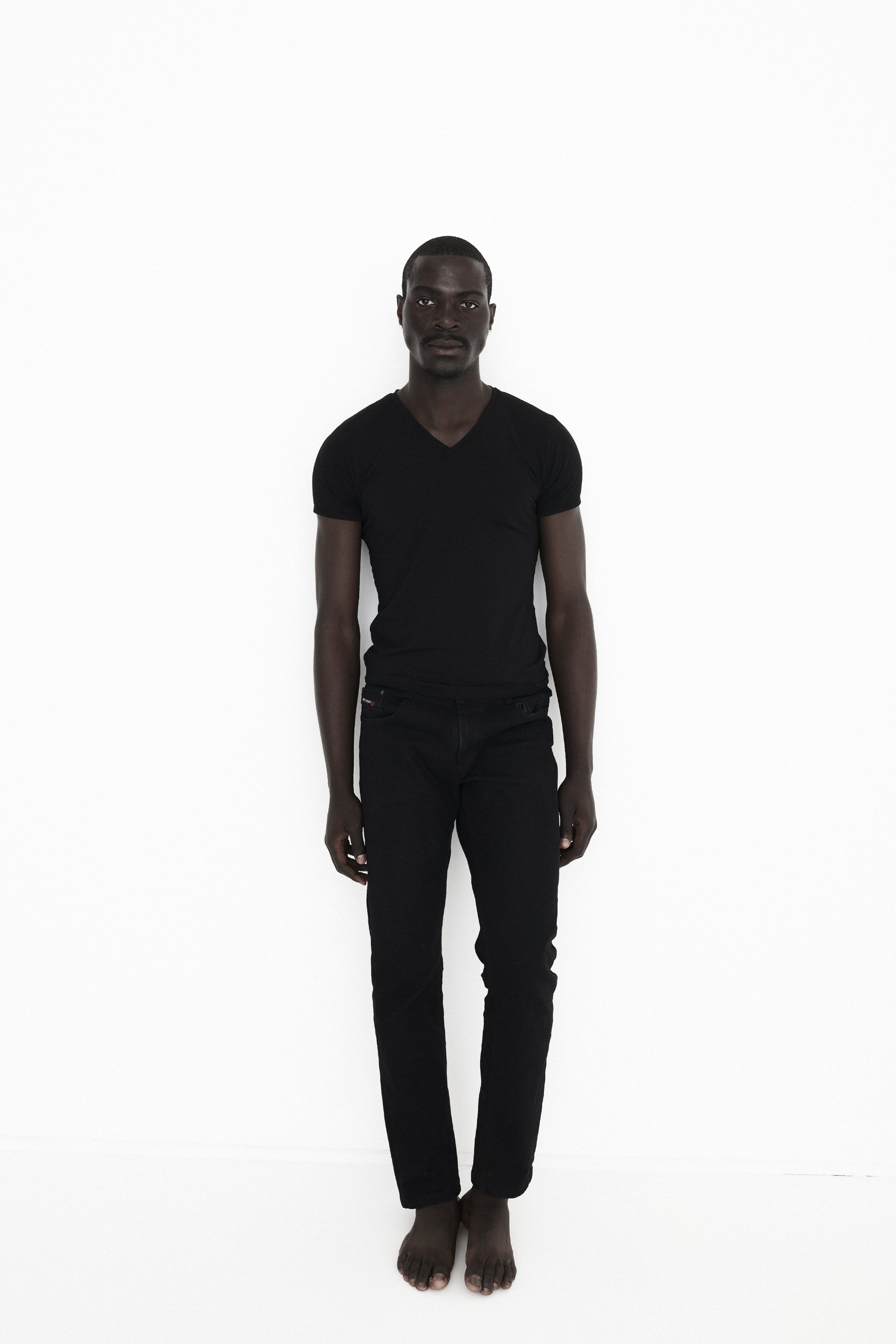 A tall man with dark skin standing against a plain white background, wearing a black t-shirt and black pants, barefoot.
