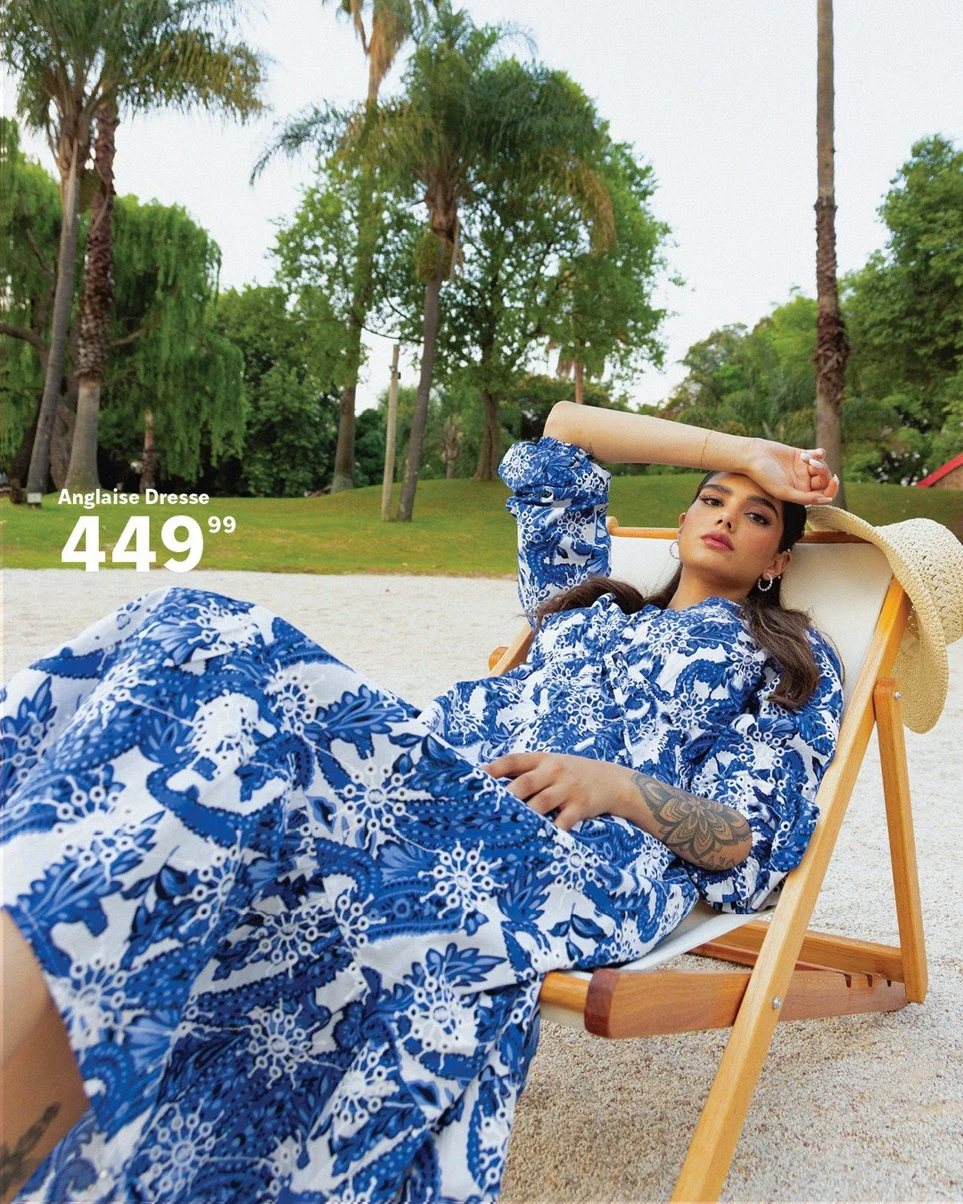 A woman lying on a lounge chair outdoors, wearing a blue and white patterned dress and a straw wide-brim hat, with trees in the background.