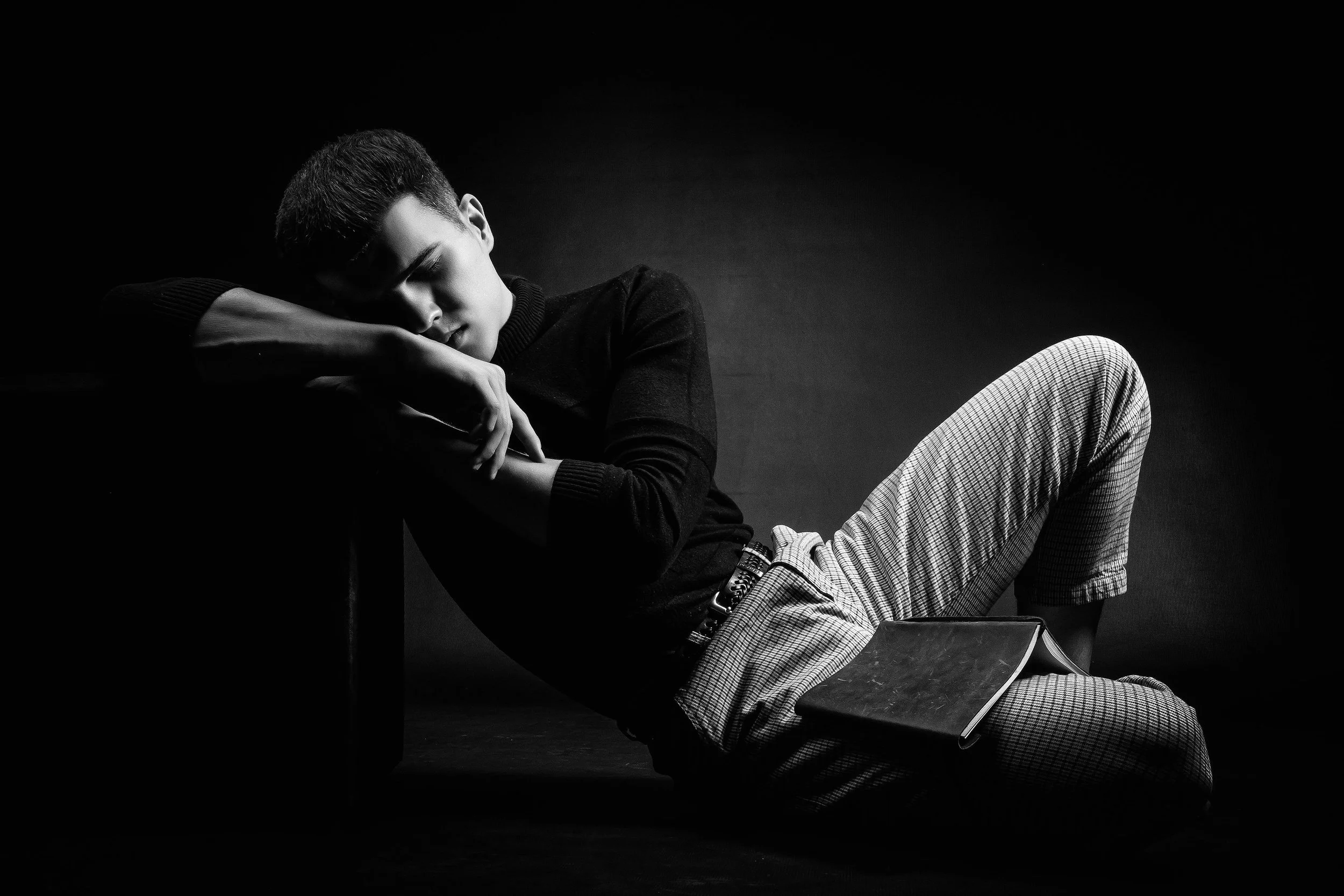A young man with short hair, wearing glasses, a dark sweater, and light checkered pants, reclines on the floor with his eyes closed, resting his head on his arm, and holding a closed book.