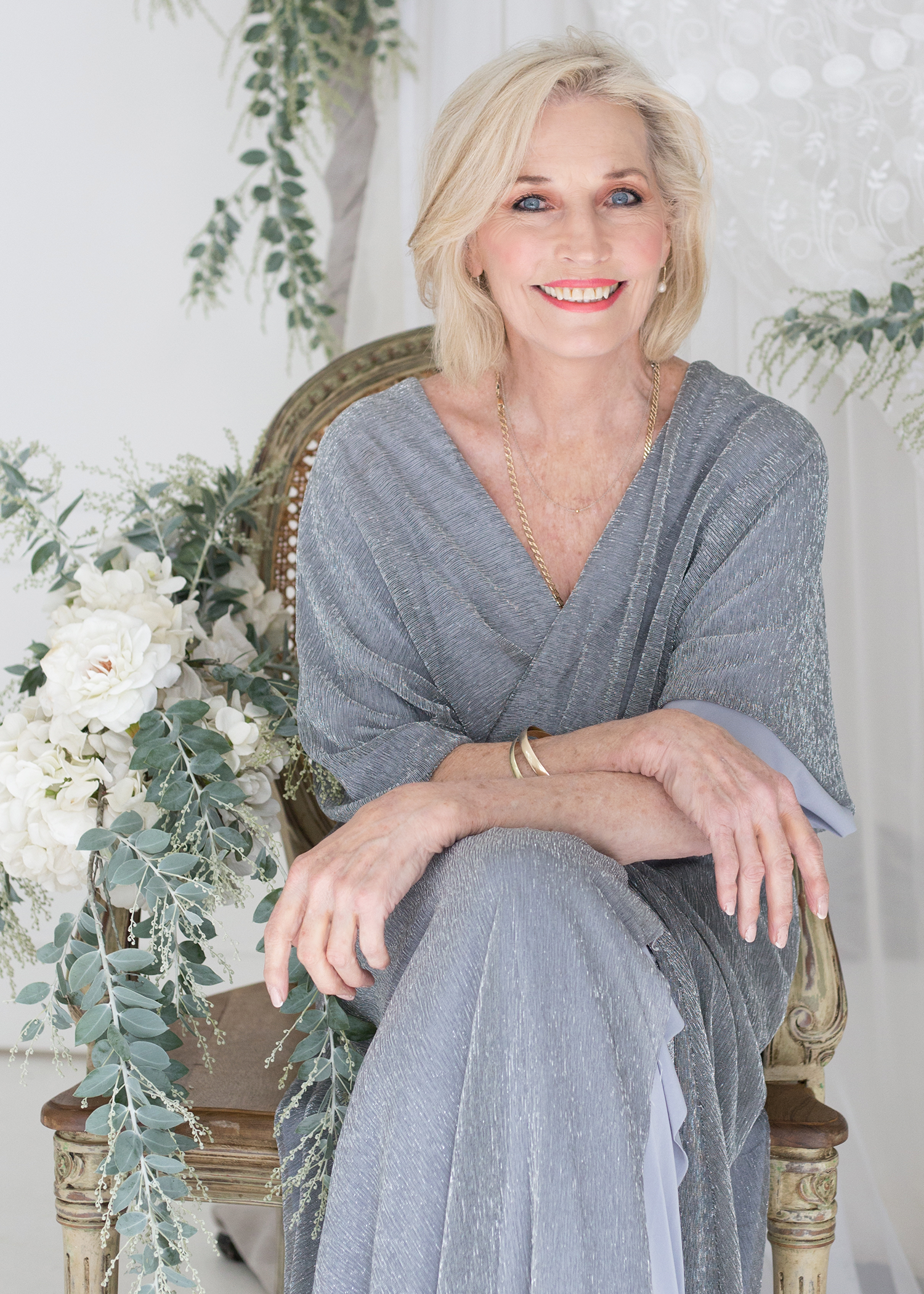 MARIETA Ferreira from AURA Models with blonde hair and blue eyes, smiling, seated on a wooden chair with floral and greenery decorations around her, wearing a silver-gray dress and jewelry.