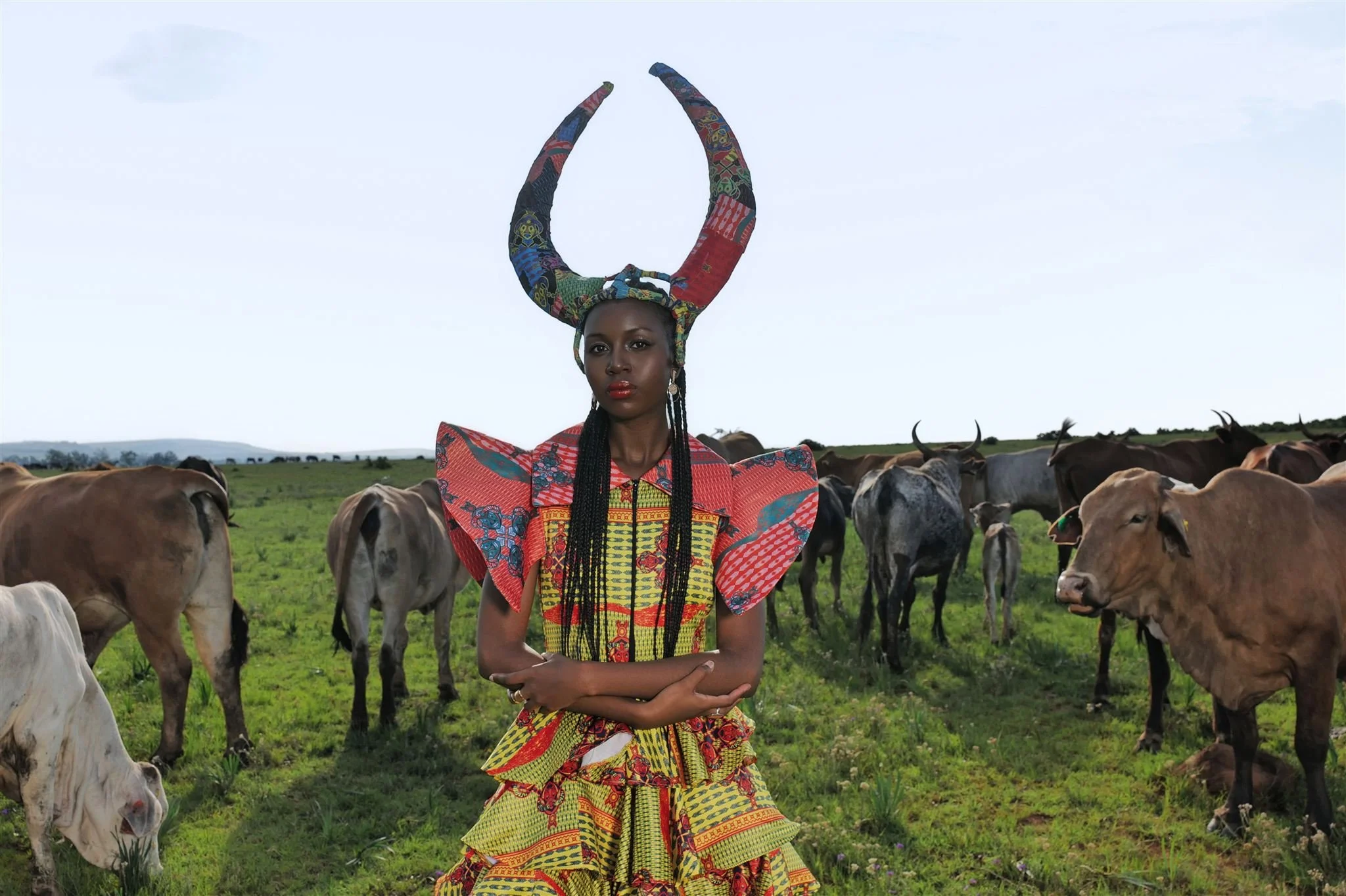 RIVO Baloyi from AURA Models in colorful traditional Masamara African clothing stands in a grassy field with cows, with a clear sky in the background.