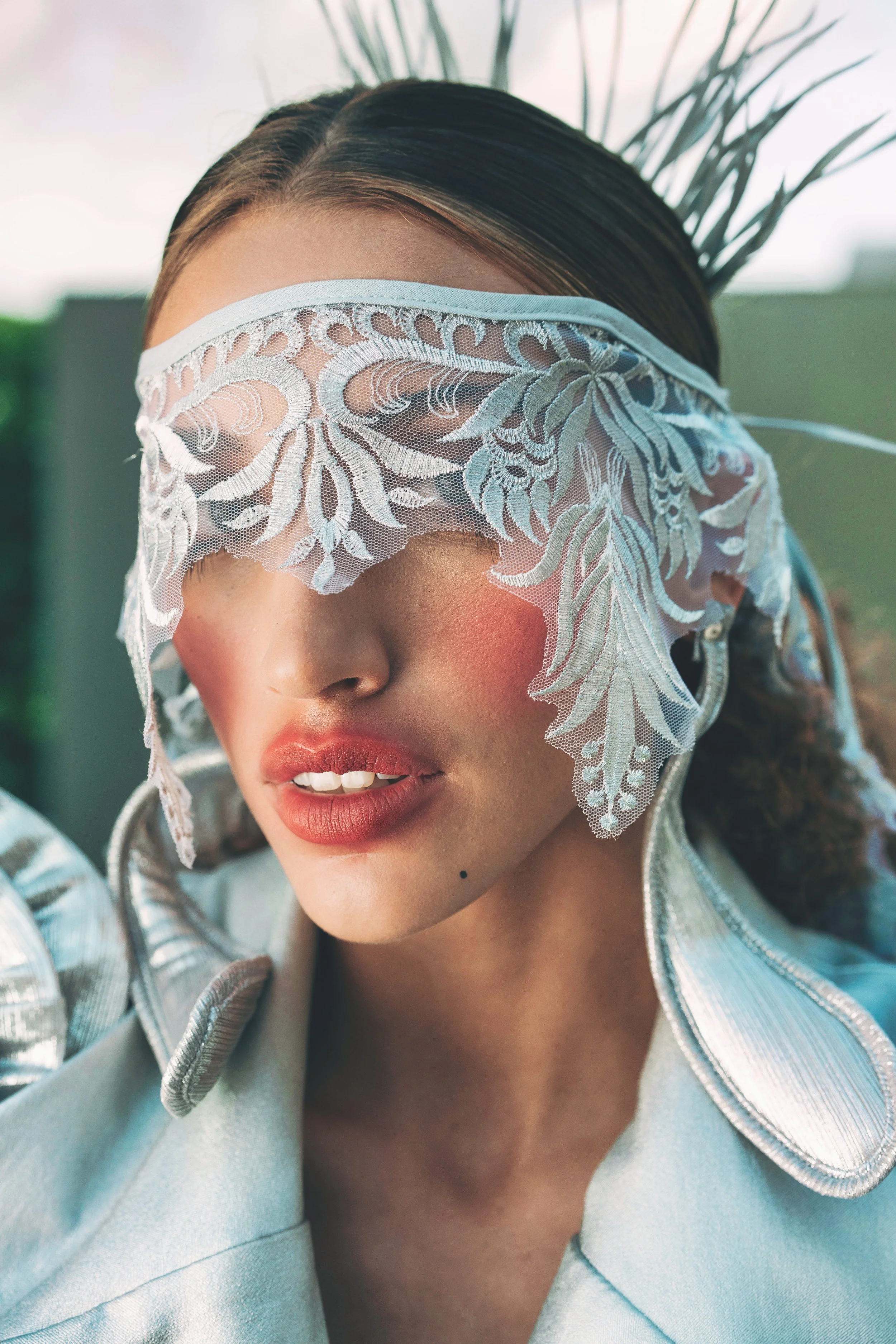 Close-up of MEGAN Leigh from AURA Models wearing a white lace eye mask and a shiny silver jacket, with a green plant in the background.