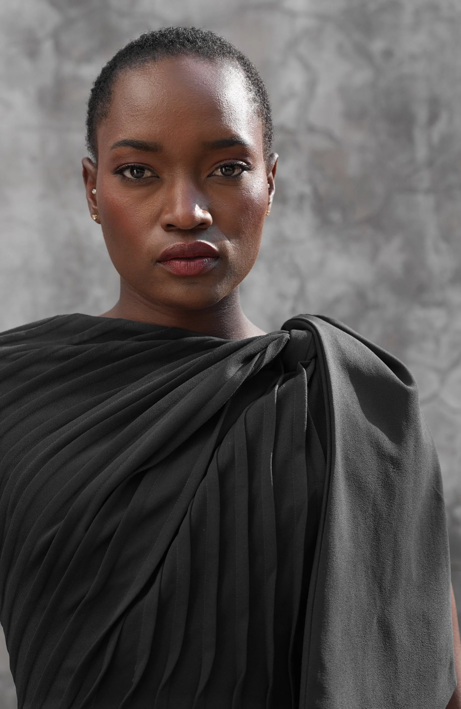 Portrait of SHALOM Musvosvi from AURA Models with short hair, wearing makeup, earrings, and a black draped garment against a gray background.