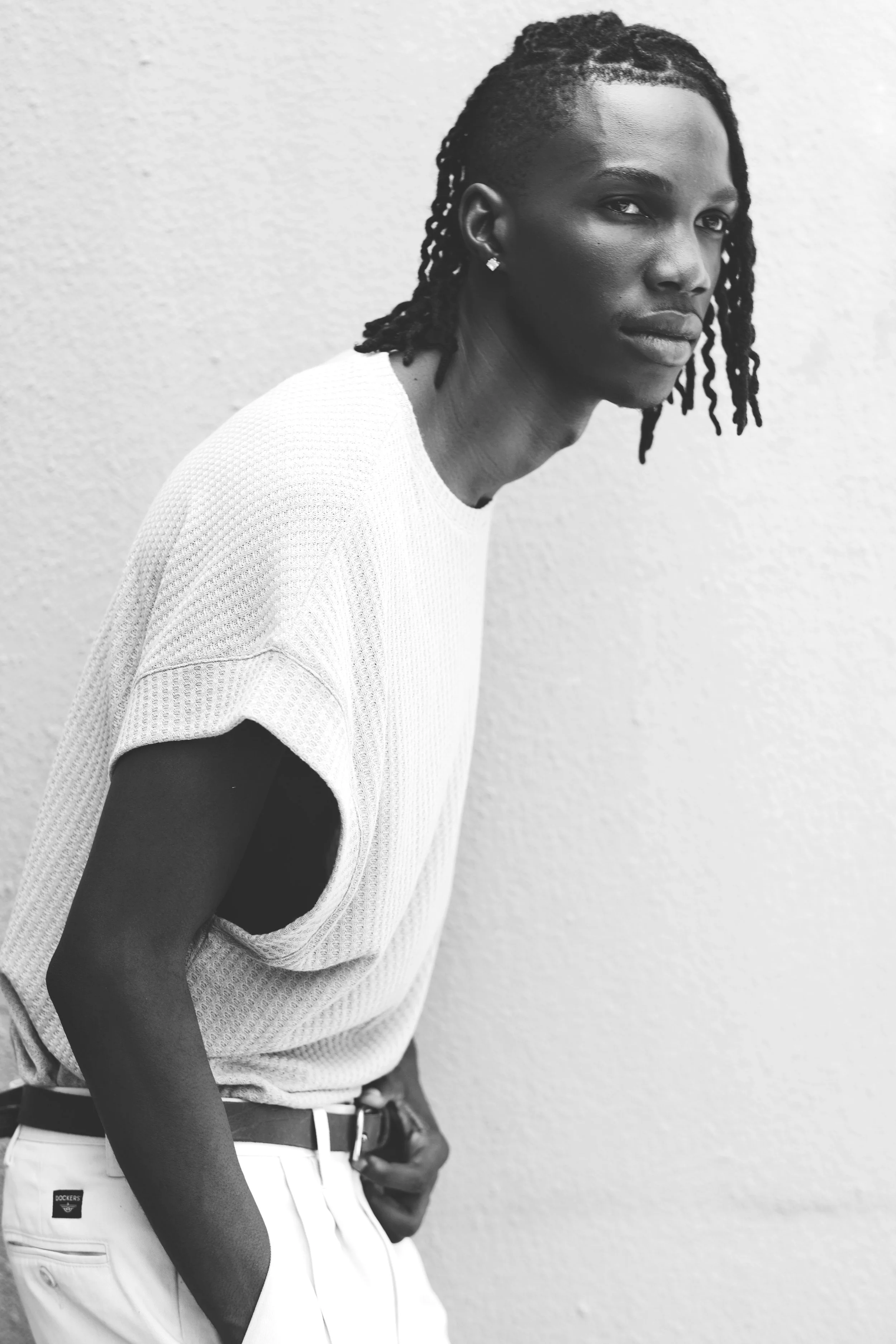A black and white photo of TSHEDZA Ramavheya from AURA Models with dreadlocks wearing a loose, textured T-shirt and light-colored pants, standing against a plain wall.