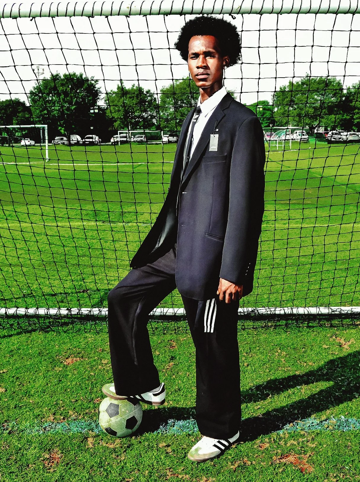FRANK Mongezi from AURA Models in a black suit and sportswear shirt, standing on a soccer field with his foot on a soccer ball, in front of a goal net, with trees and parked cars in the background.