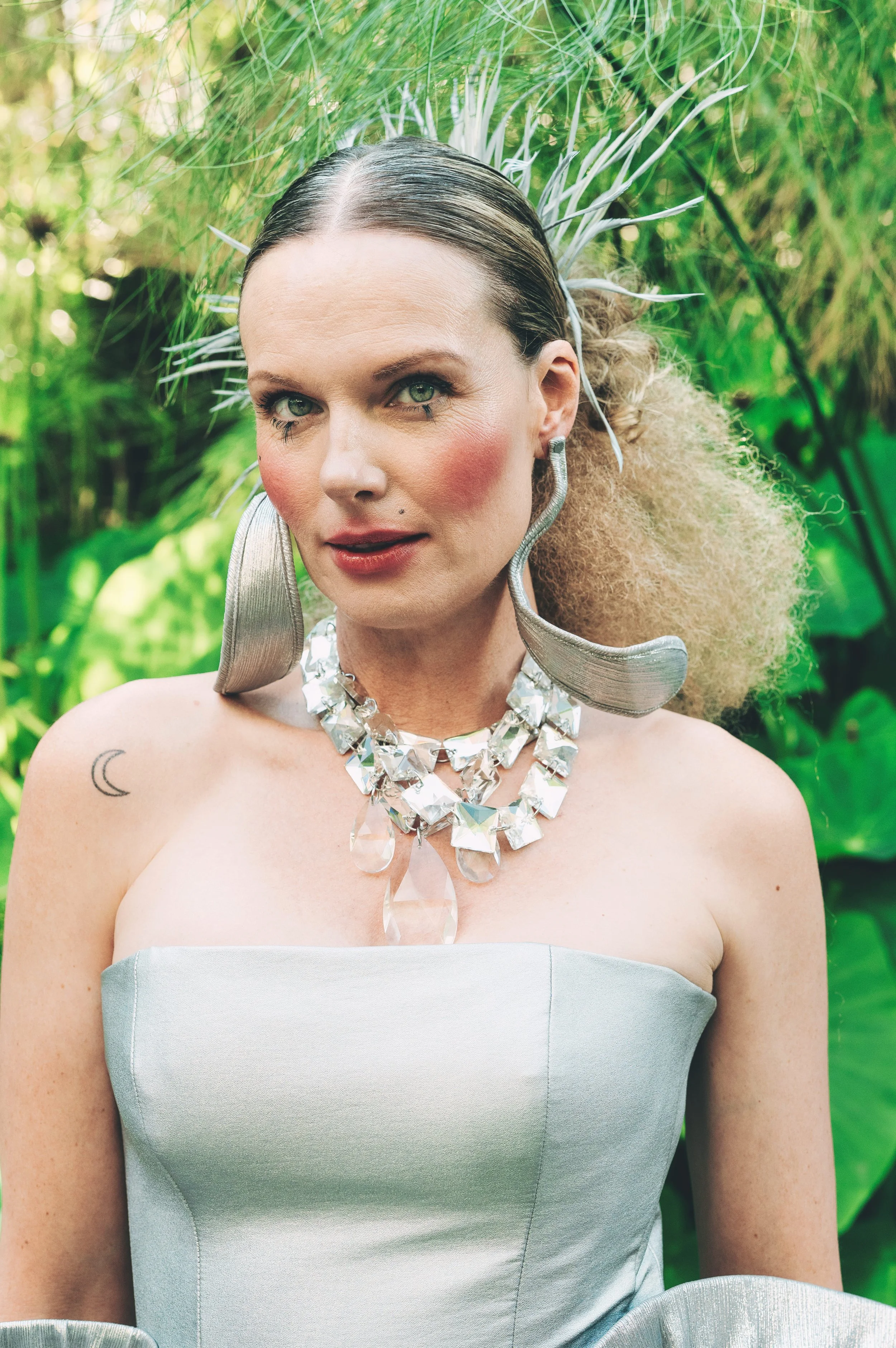 NATALIA Kostanova from AURA Models with green eyes and light skin draped in a silver-grey strapless gown, standing outdoors with lush green foliage in the background. 