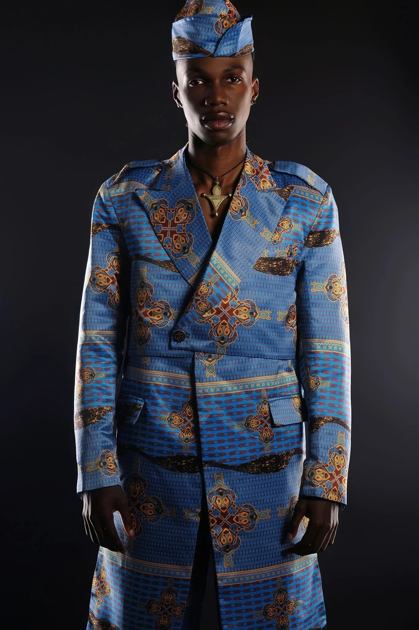 TSHEDZA Ramavheya from AURA Models wearing a matching Masamara blue patterned suit with intricate designs, complemented by a matching headscarf, against a dark background.