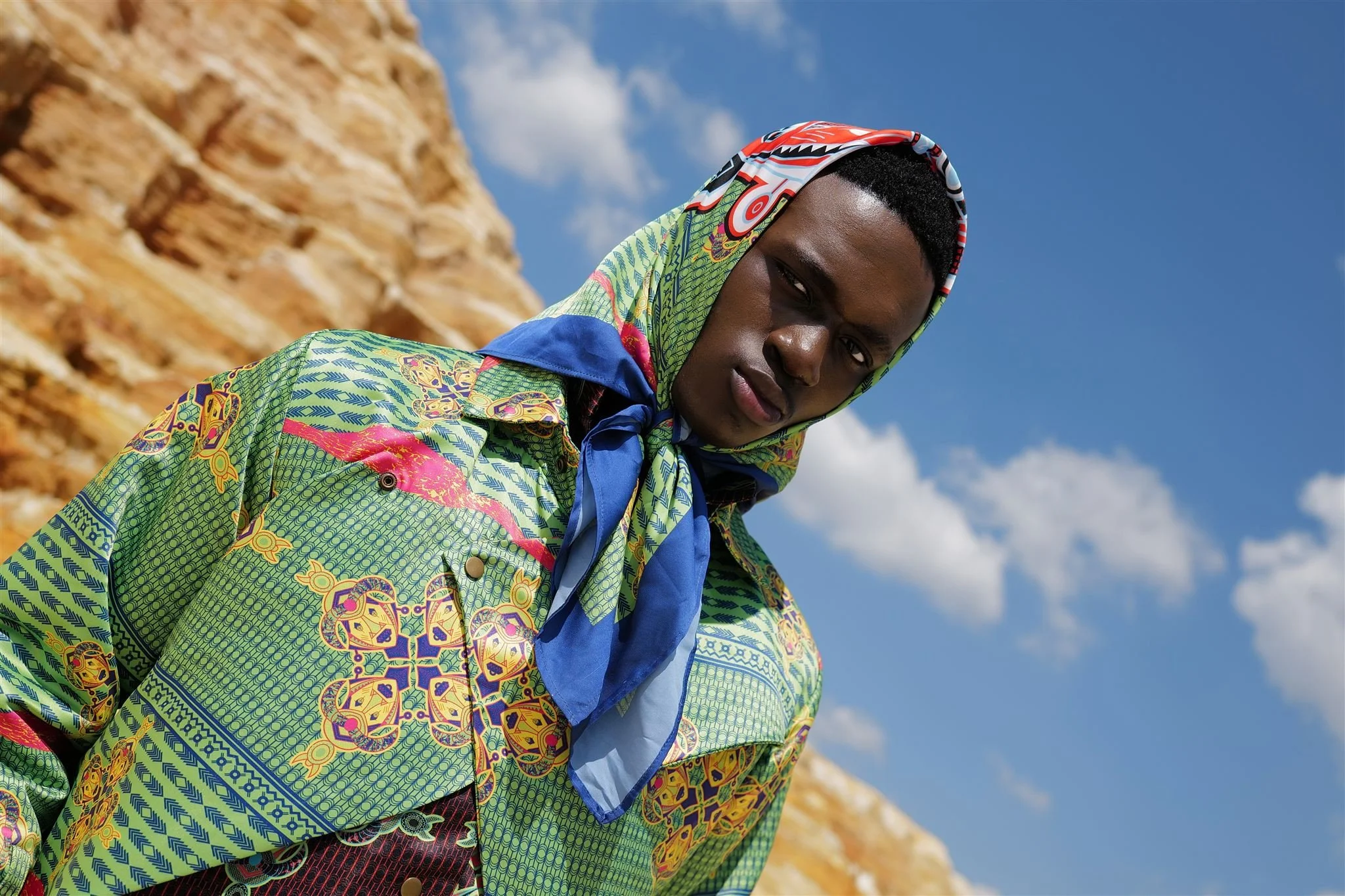 MBHURI Baloyi from AURA Models wearing a colorful patterned Masamara shirt and a blue headscarf outdoors against a rocky landscape and blue sky with clouds.
