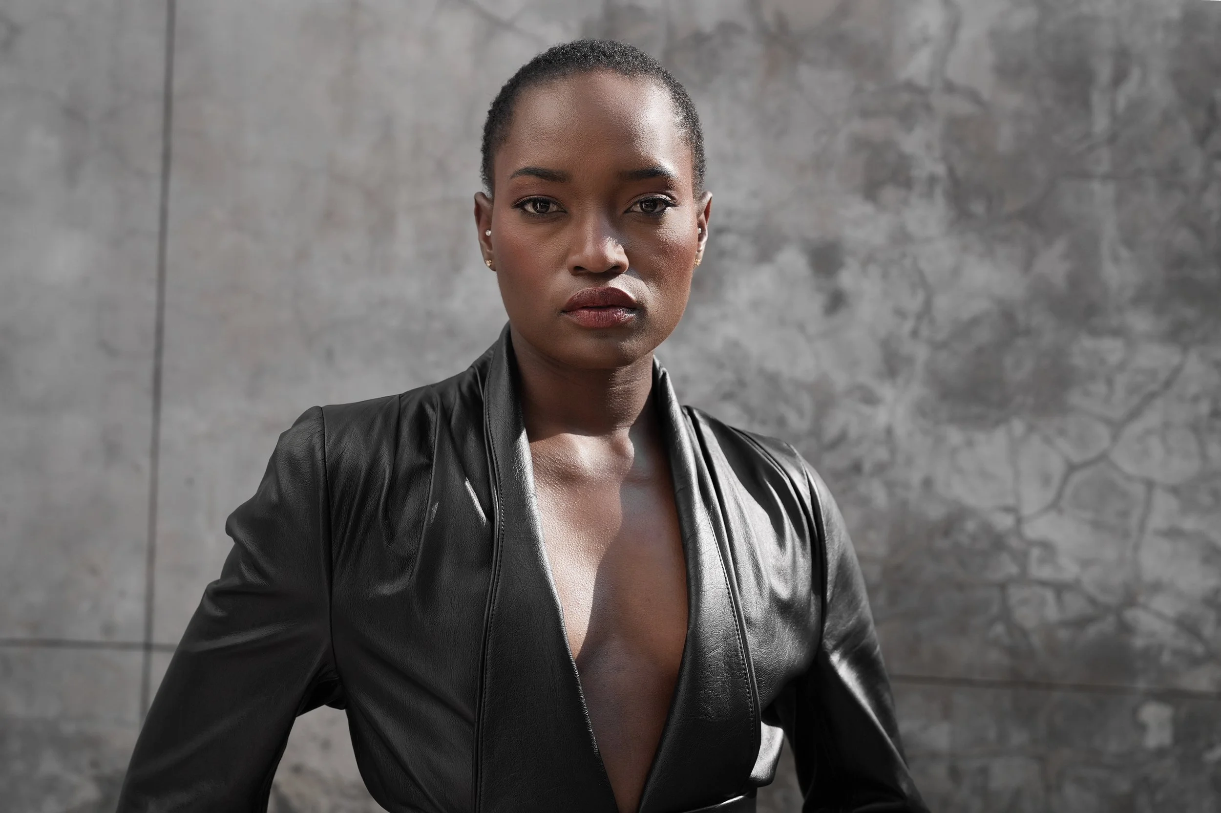 A confident SHALOM Musvosvi from AURA Models with short hair, wearing a black leather jacket, standing against a textured gray wall.