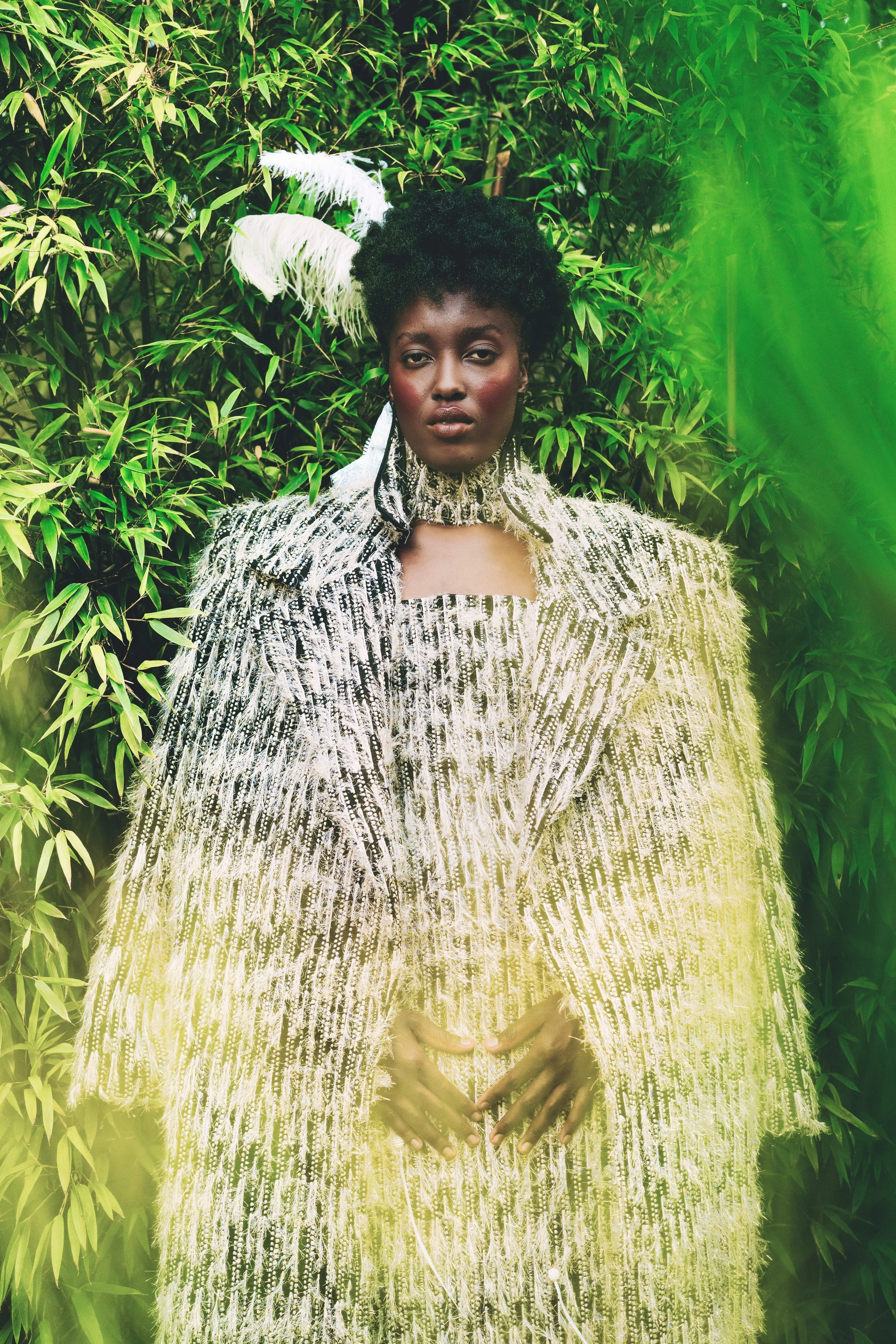 KIM Chakanyuka from AURA Models with dark skin and natural black hair wearing a silver ornate dress with feathered embellishments, standing in front of dense green foliage, with a glowing green light effect on the right side.